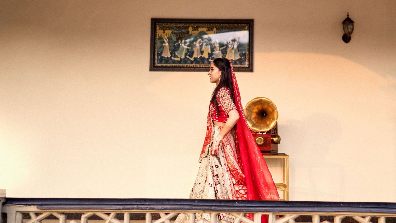 Woman in red and gold Indian bridal lehenga standing on balcony beside vintage gramophone with ornate painting.