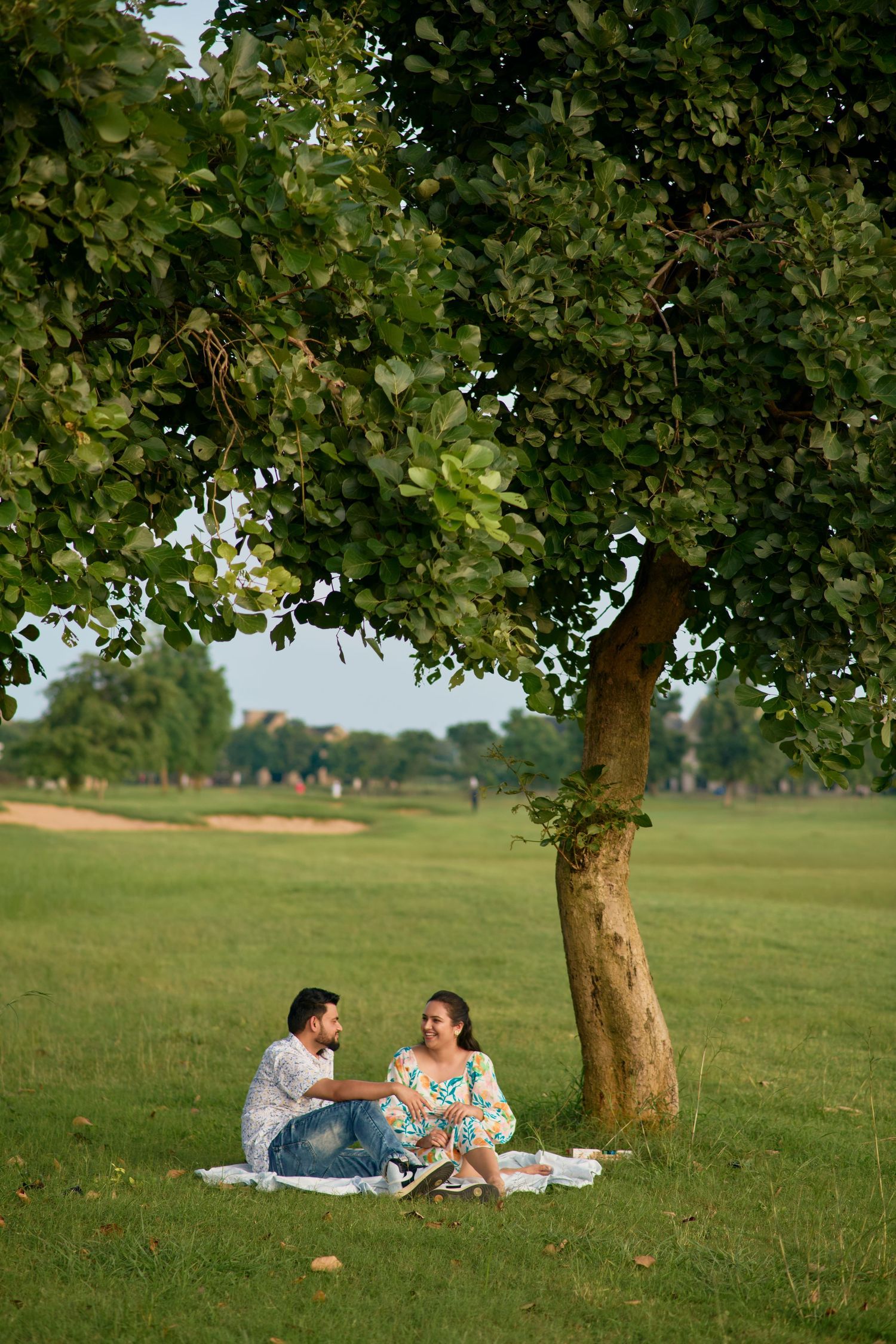 Couple sitting on a blanket under a large tree in a lush green park, enjoying a romantic outdoor picnic together.