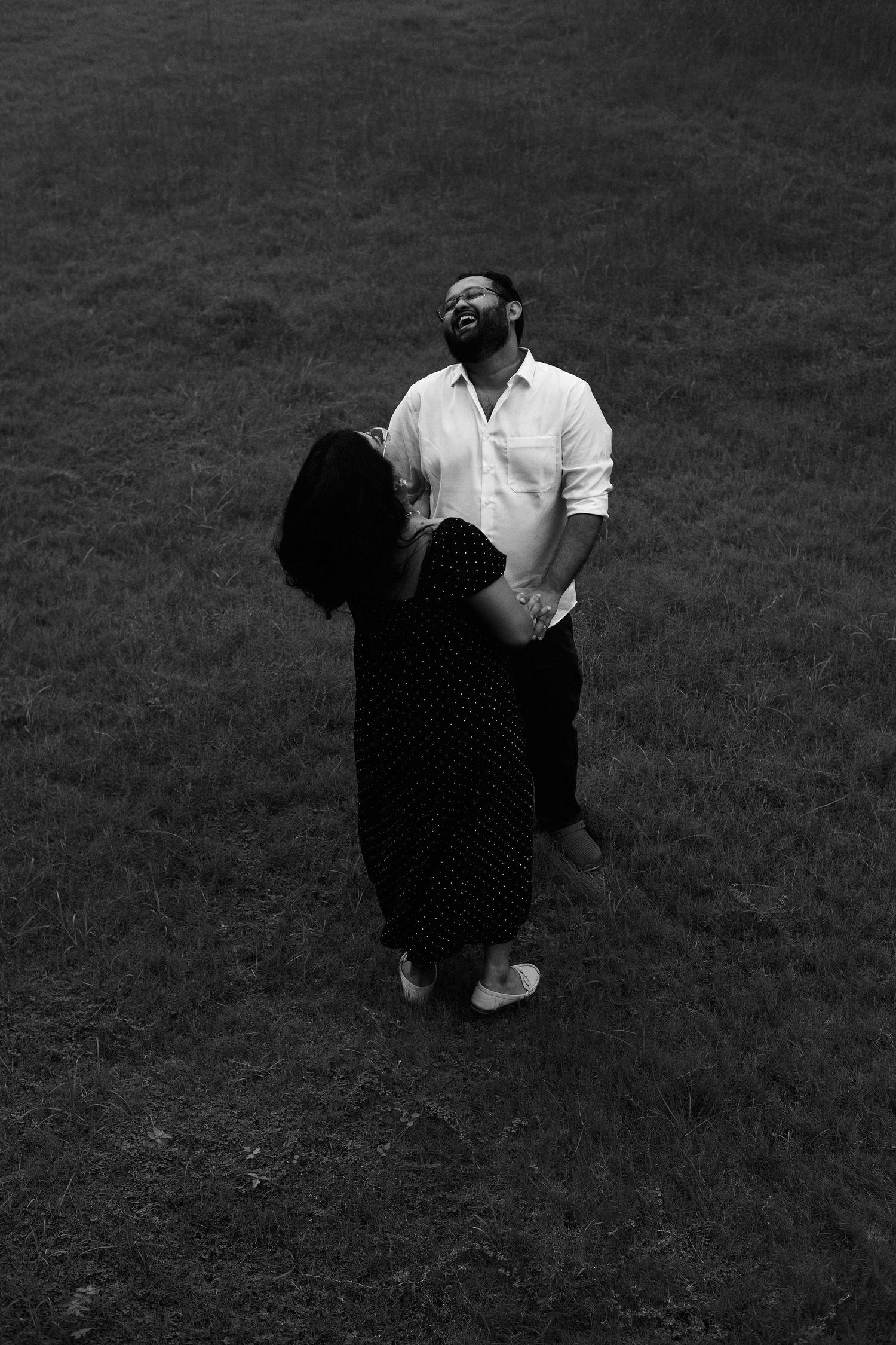Black and white photo of a couple embracing and laughing together in a grassy field outdoors.