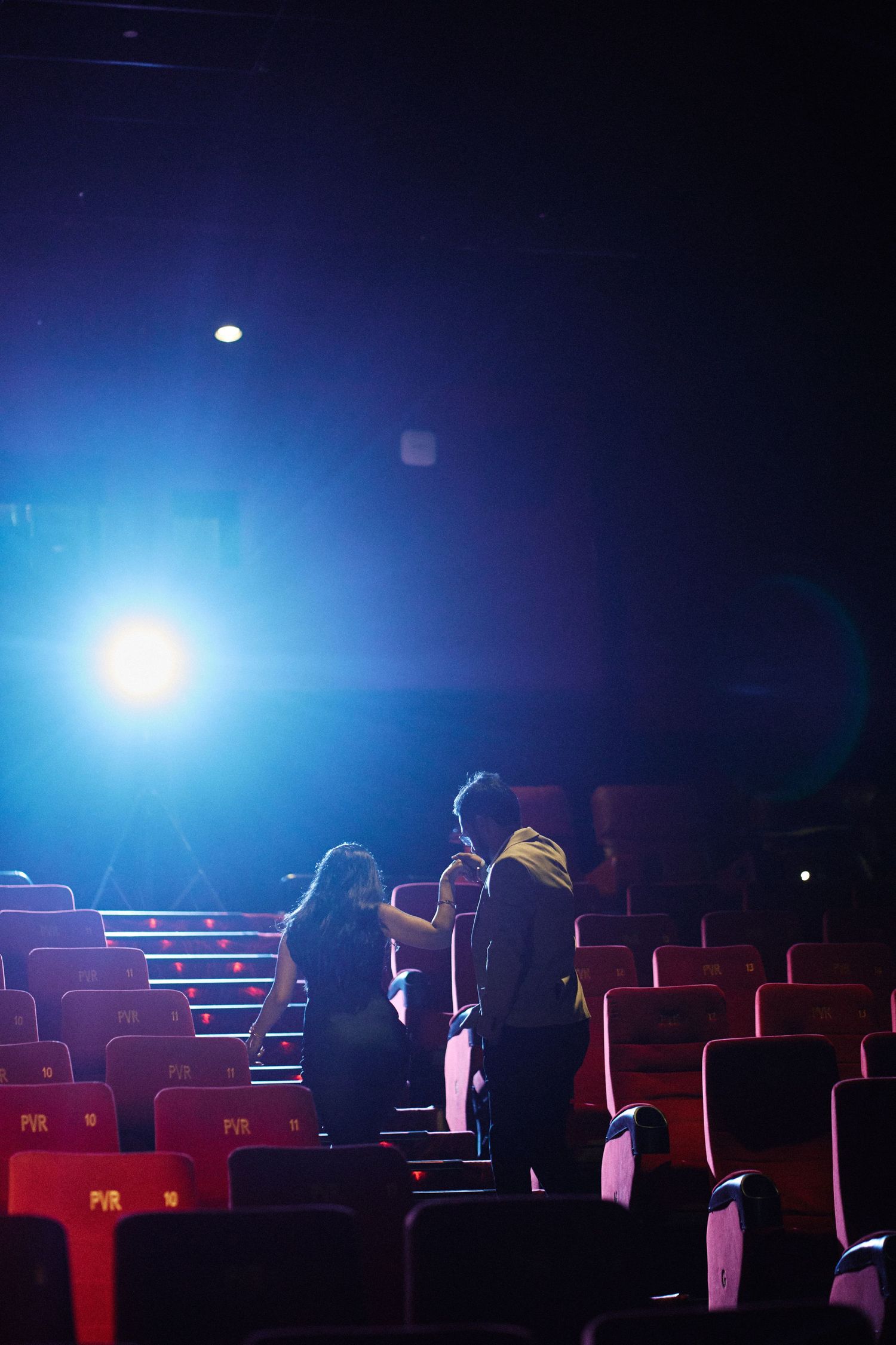Two people standing in an empty red-seated cinema hall with dramatic blue stage lighting.