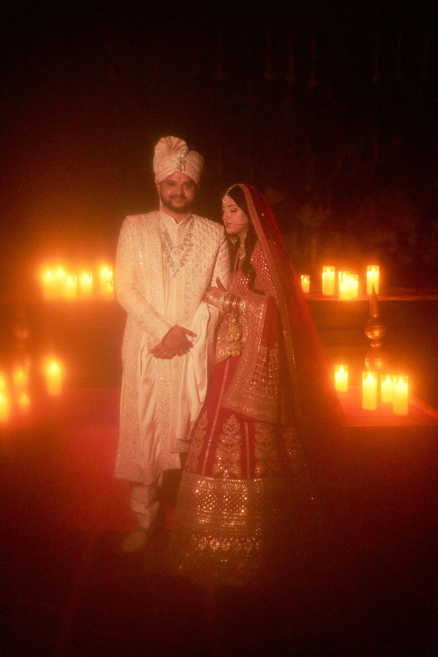 Indian bride and groom in traditional attire surrounded by glowing candles in romantic setting.