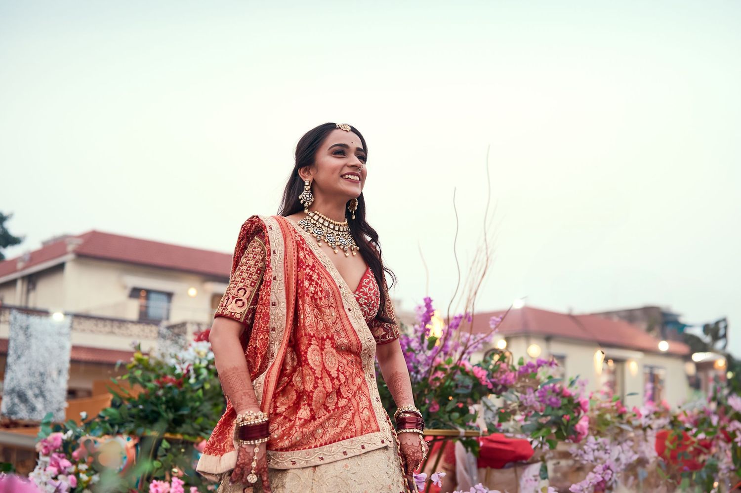 Bride in red and gold lehenga with jewelry smiling outdoors among colorful flowers at an Indian wedding.