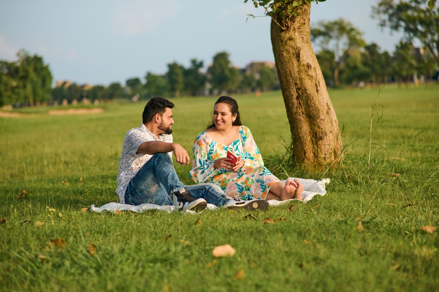 Couple sitting on blanket in green park near tree, enjoying romantic outdoor picnic on sunny day.