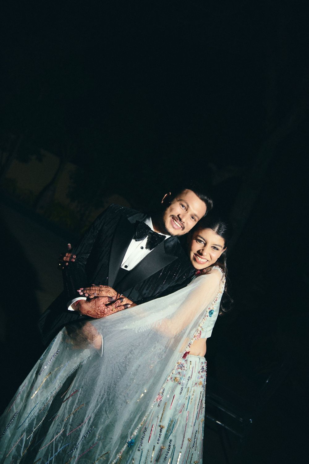 Couple posing elegantly, man in black tuxedo and woman in light blue embroidered gown with henna hands.