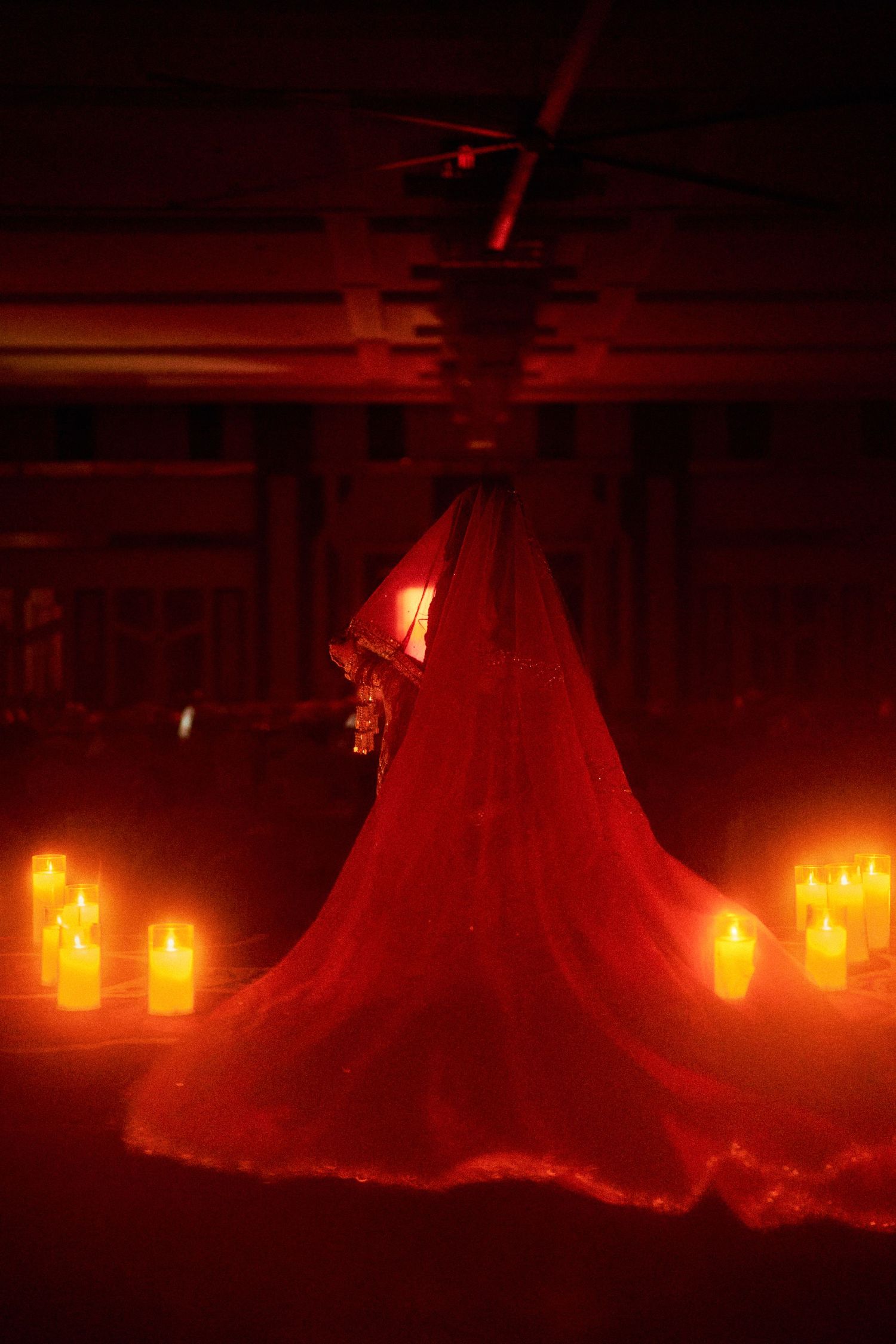 Bride in red gown surrounded by glowing candles in dramatic red-lit venue.