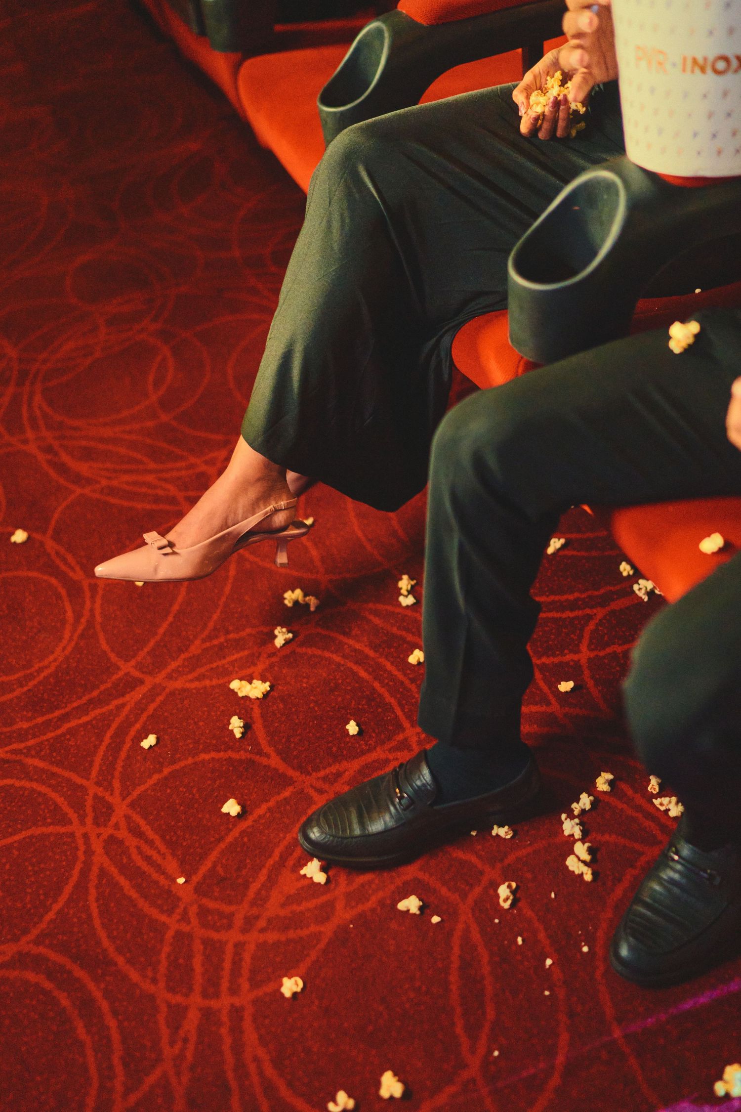 Two people sitting in cinema seats with popcorn scattered on red patterned carpet floor.