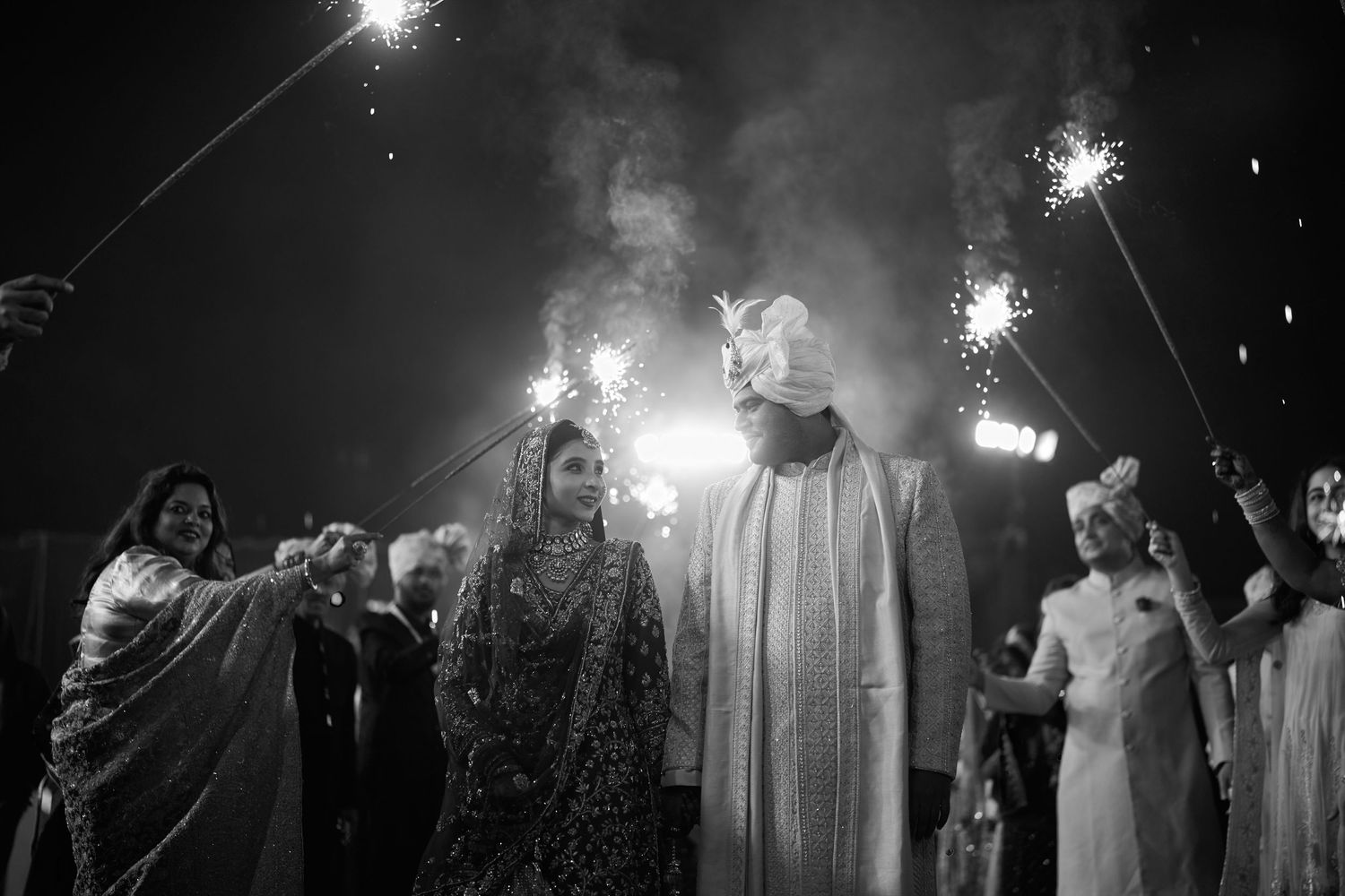 Black and white photo of Indian wedding couple walking through sparklers at night.