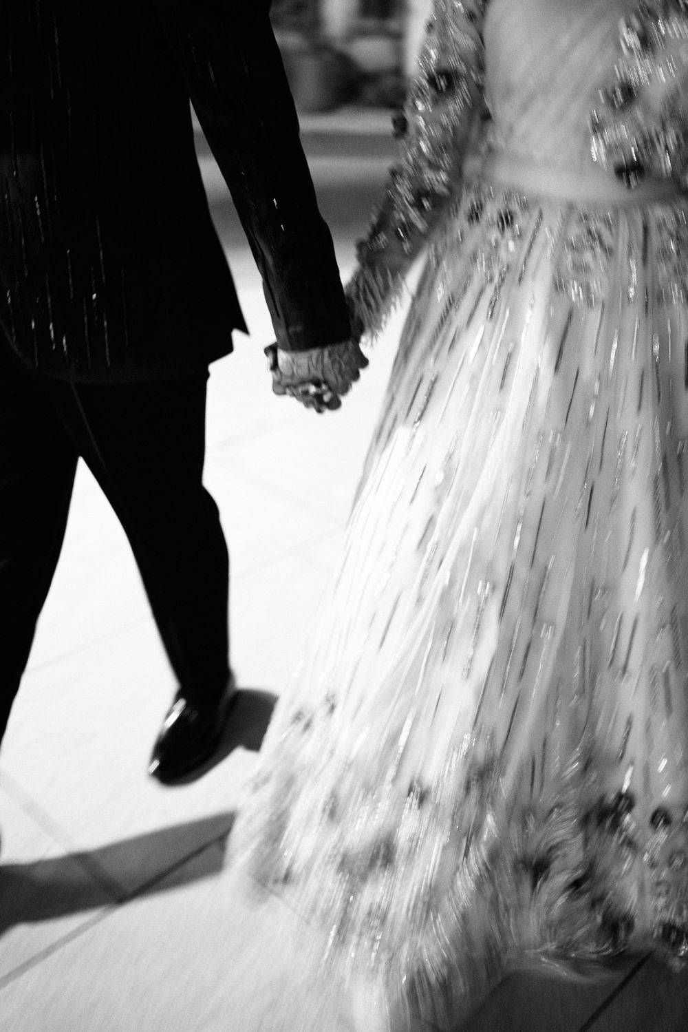 Black and white photo of a couple holding hands, bride in flowing gown and groom in suit, walking together.
