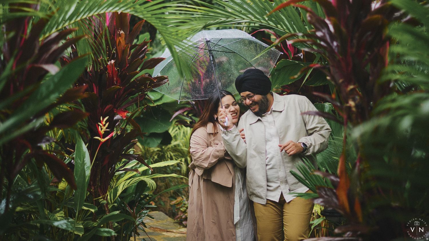 Couple sharing umbrella surrounded by lush tropical plants in a romantic outdoor garden photoshoot.