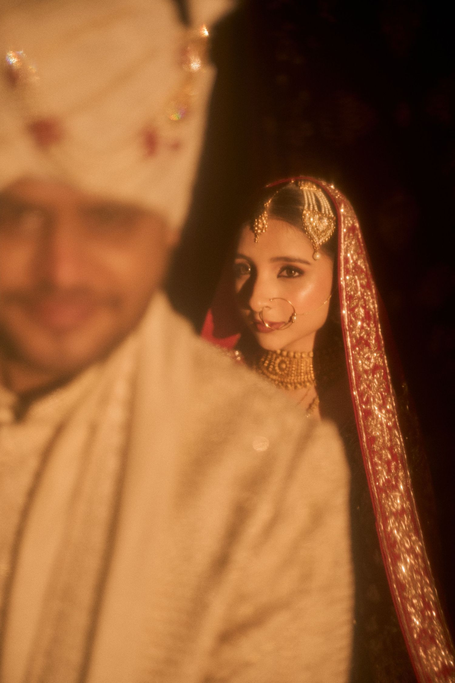 Indian bride in red dupatta and gold jewelry smiles behind groom in white sherwani and turban at wedding.