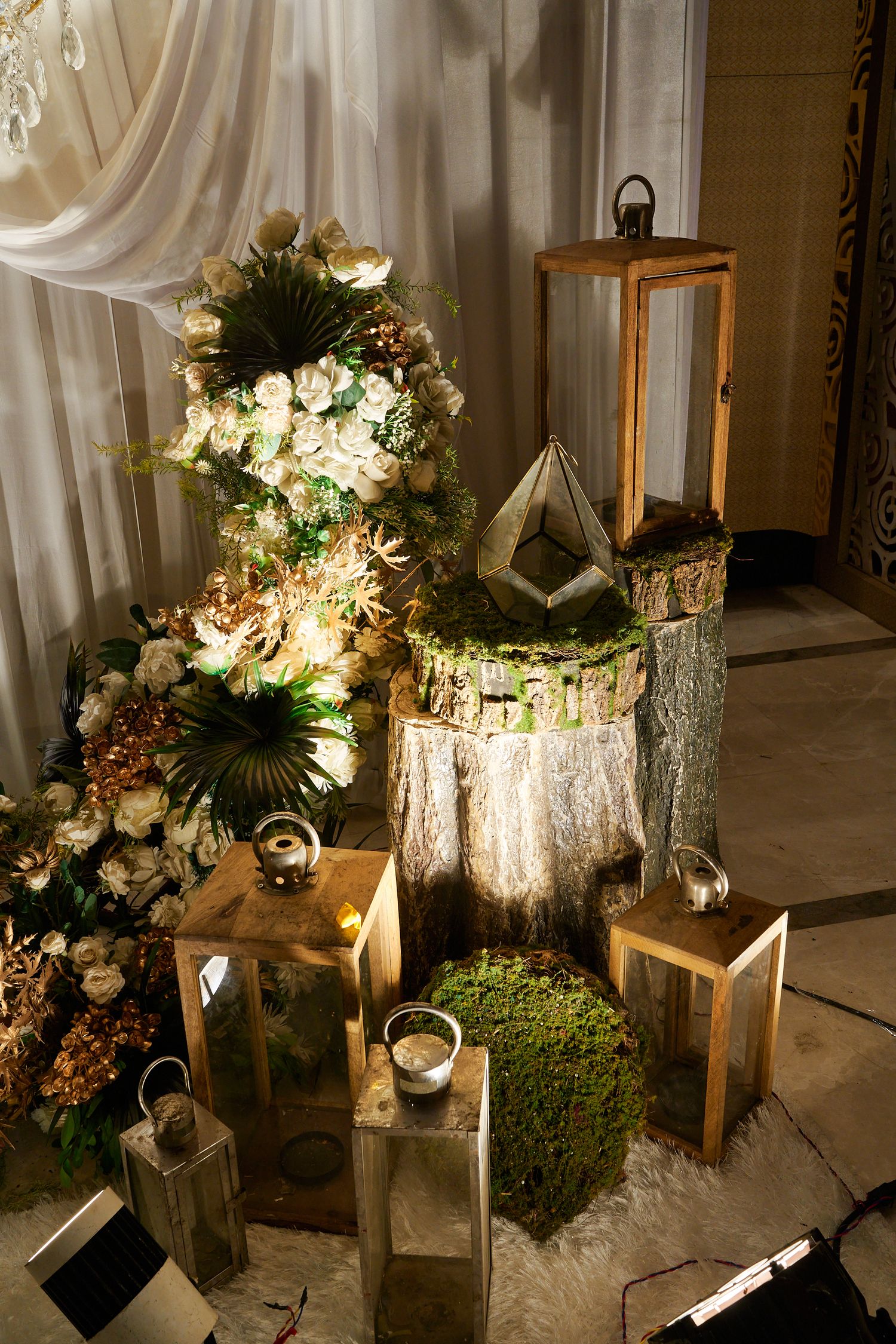Enchanting wedding decor with wooden lanterns, moss cubes, white roses, and tropical foliage arrangement.