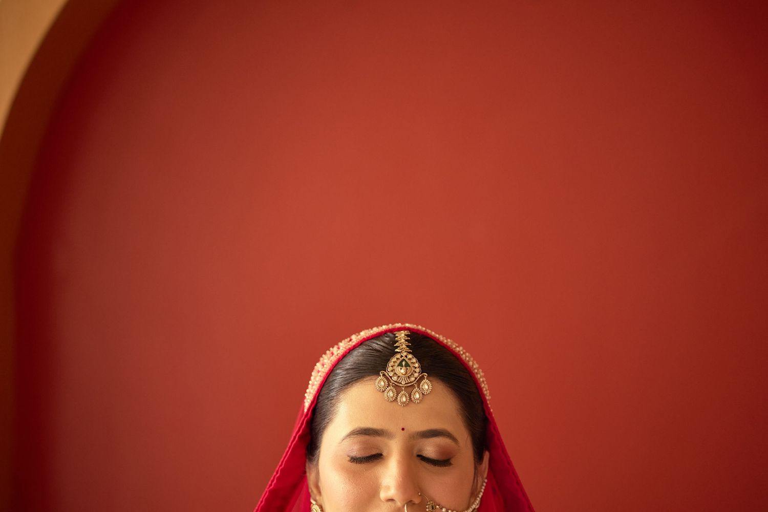 Indian bride in red dupatta with gold maang tikka jewelry against red wall.