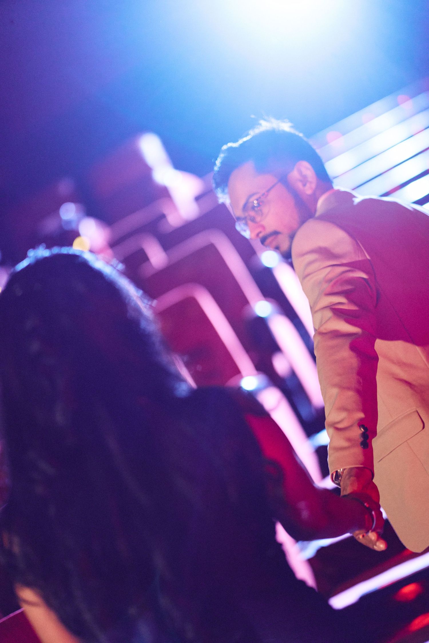 Man in white blazer and glasses at a vibrant nightclub with colorful neon lights in the background.