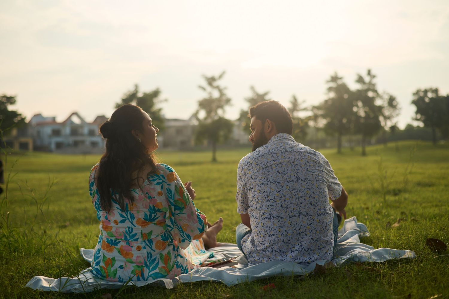 Couple sitting on blanket in green park at sunset, viewed from behind, enjoying peaceful outdoor conversation.