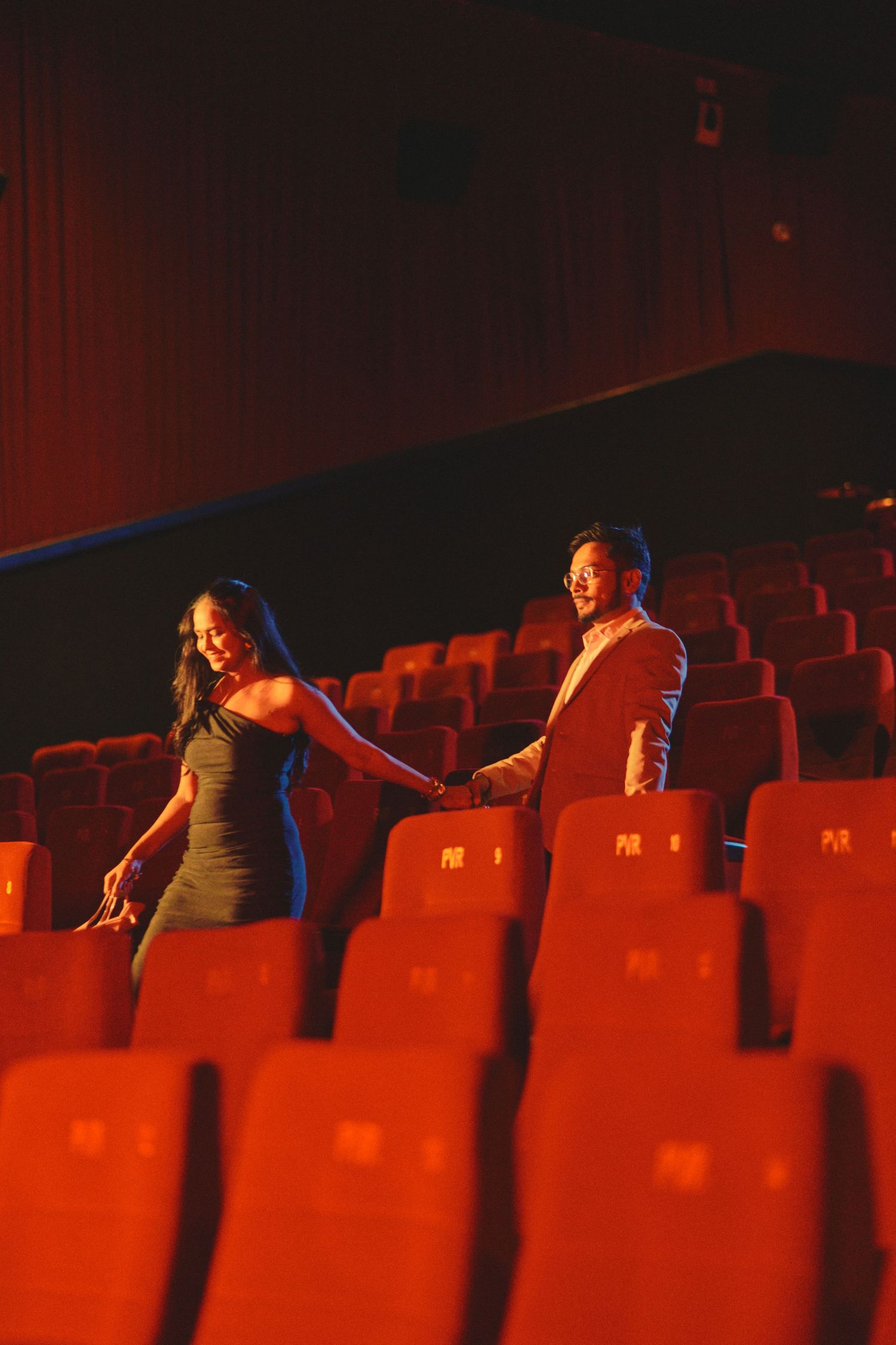 Couple holding hands in an empty red theater, dressed elegantly, sharing a romantic moment among rows of seats.