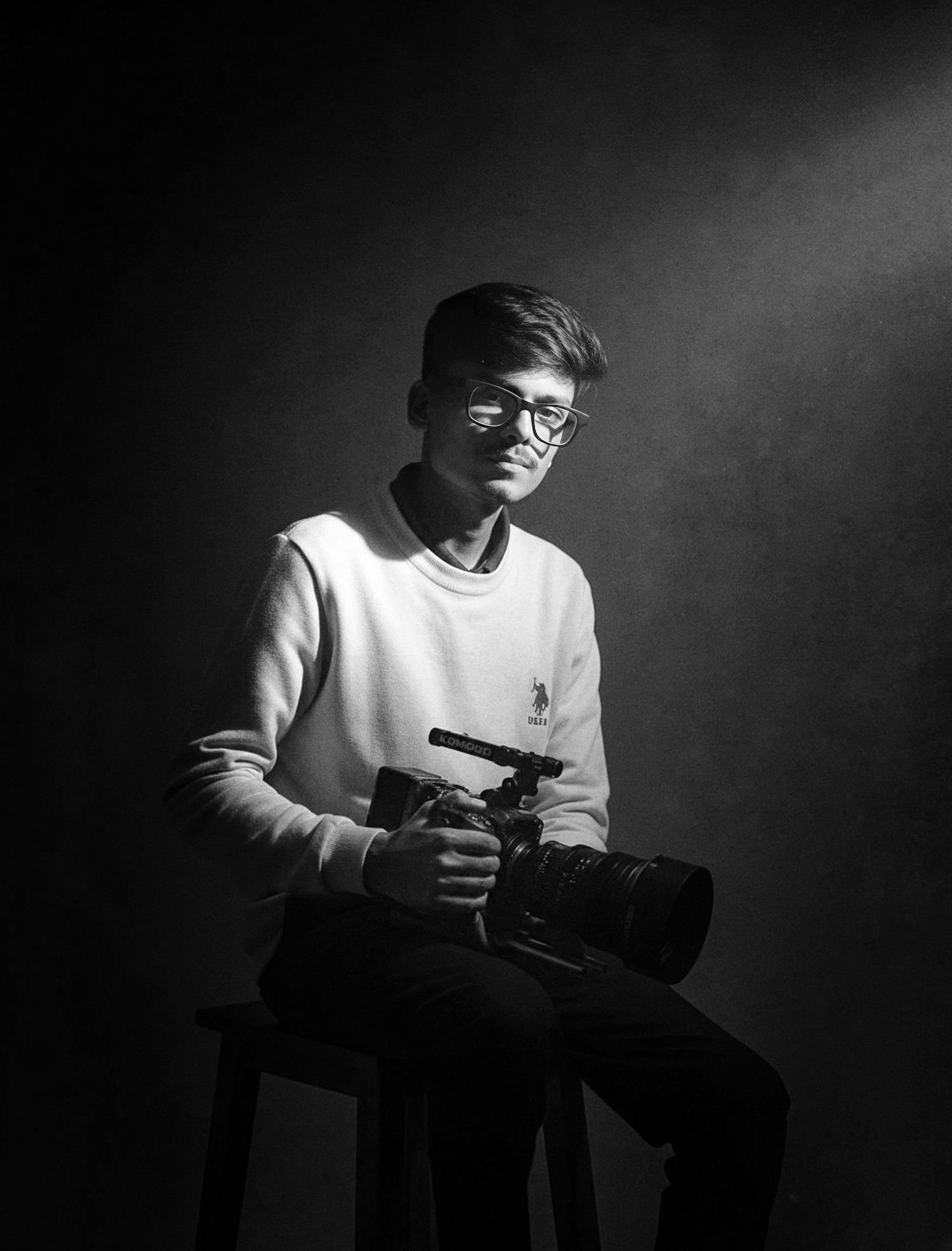Young male photographer sits on stool holding camera in dramatic black and white studio portrait.