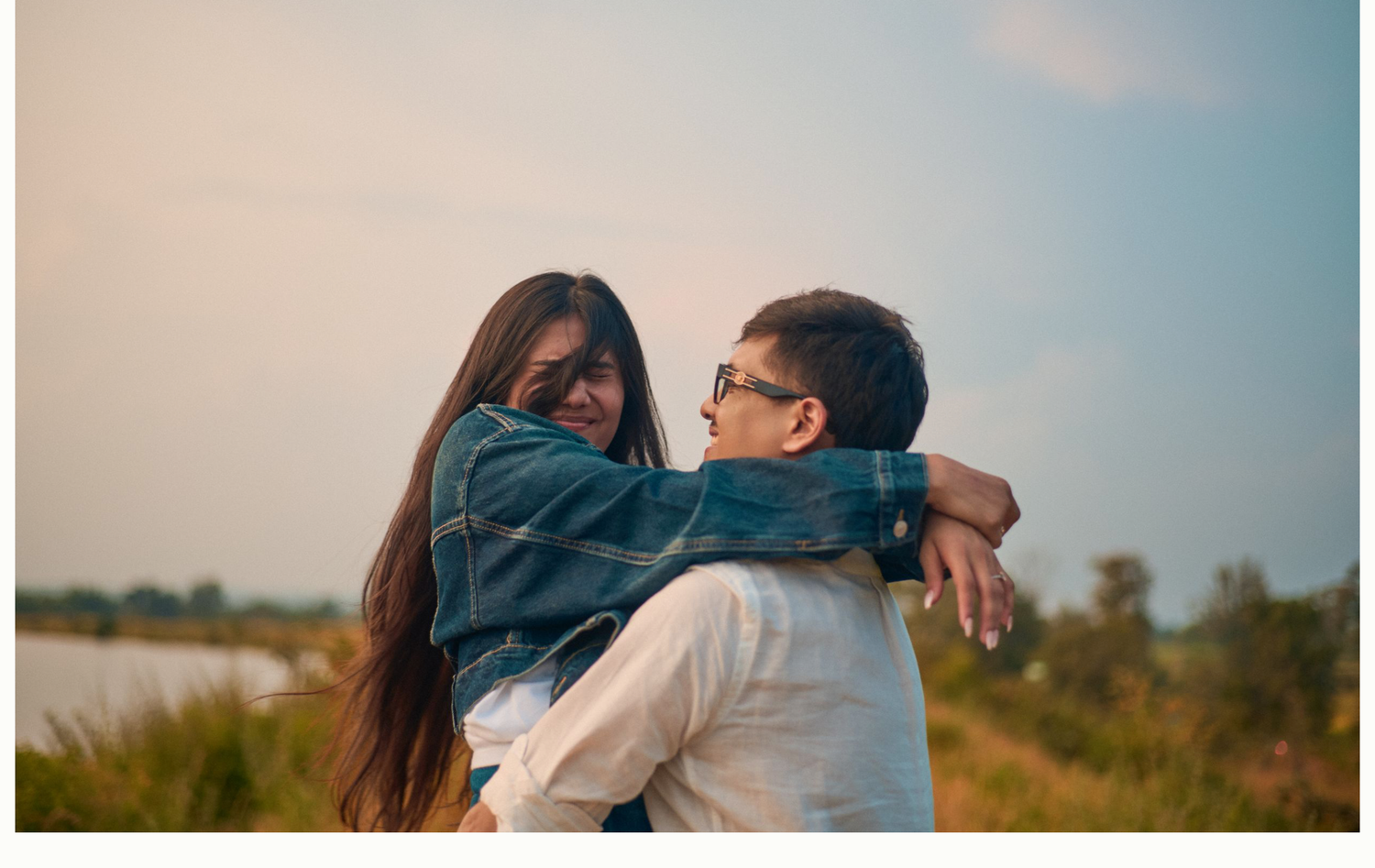 A young couple shares a romantic embrace outdoors near a lake at sunset, woman wearing denim jacket.