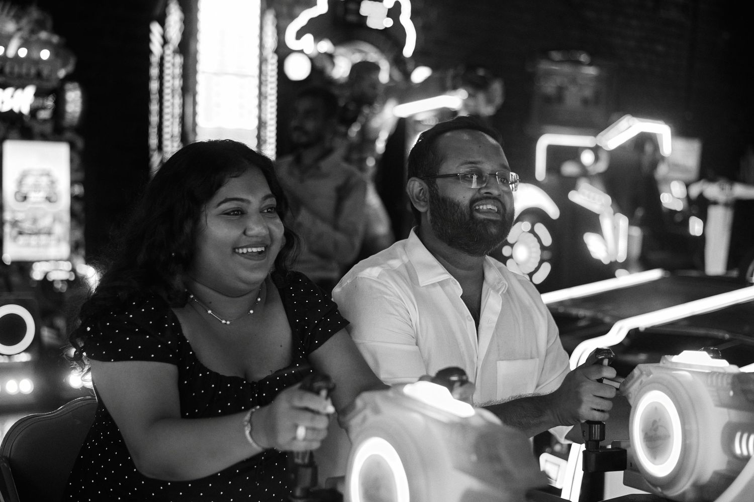 A joyful couple riding arcade bikes at a neon-lit amusement venue, captured in black and white photography.