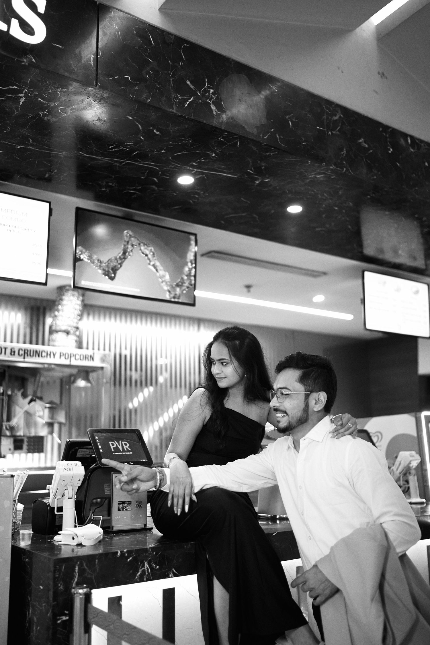 Black and white photo of a smiling couple at a PVR cinema counter, woman in black dress leaning on mans shoulder.