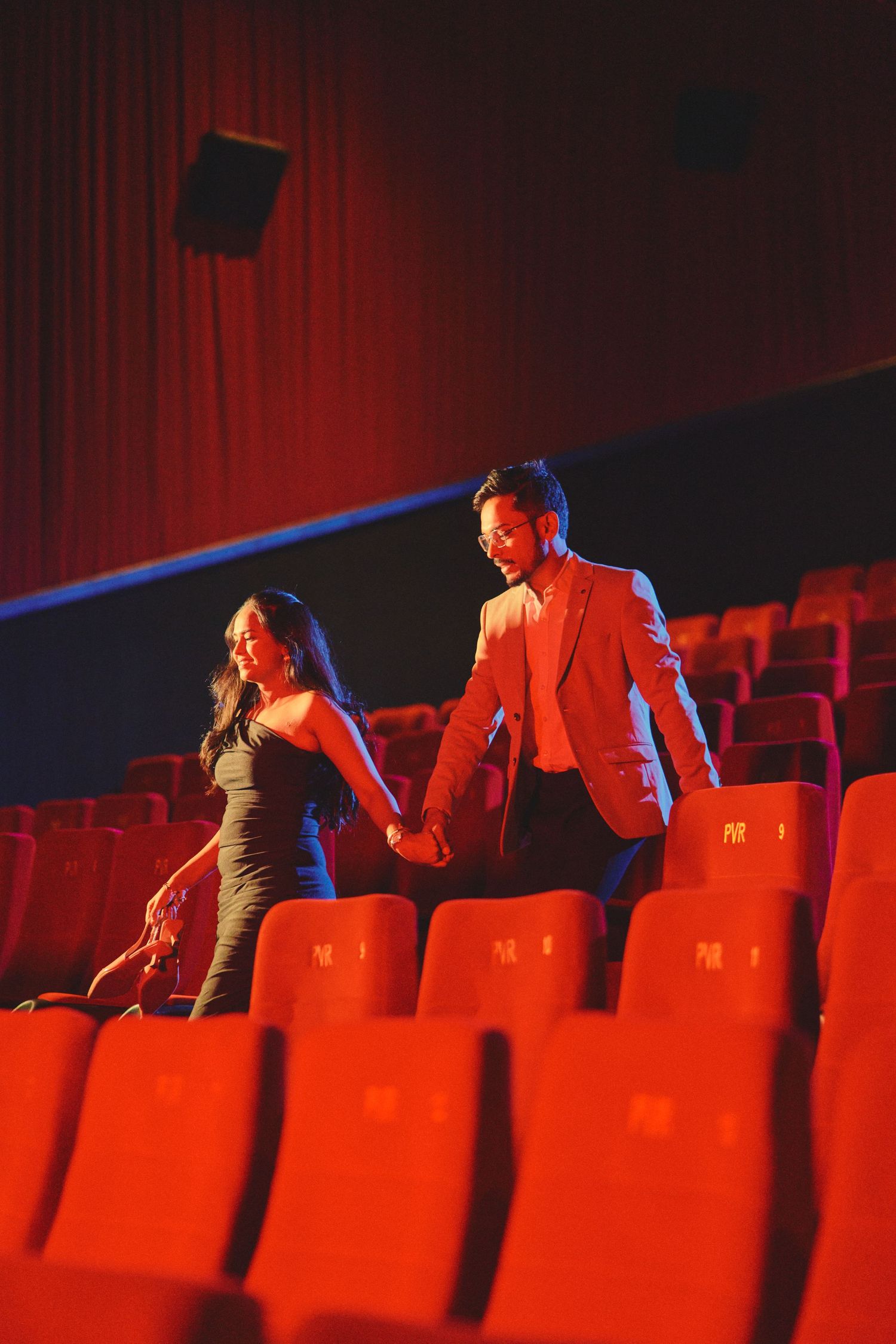 Couple holding hands in red theater seats, dressed elegantly, sharing a romantic moment in a cinema.