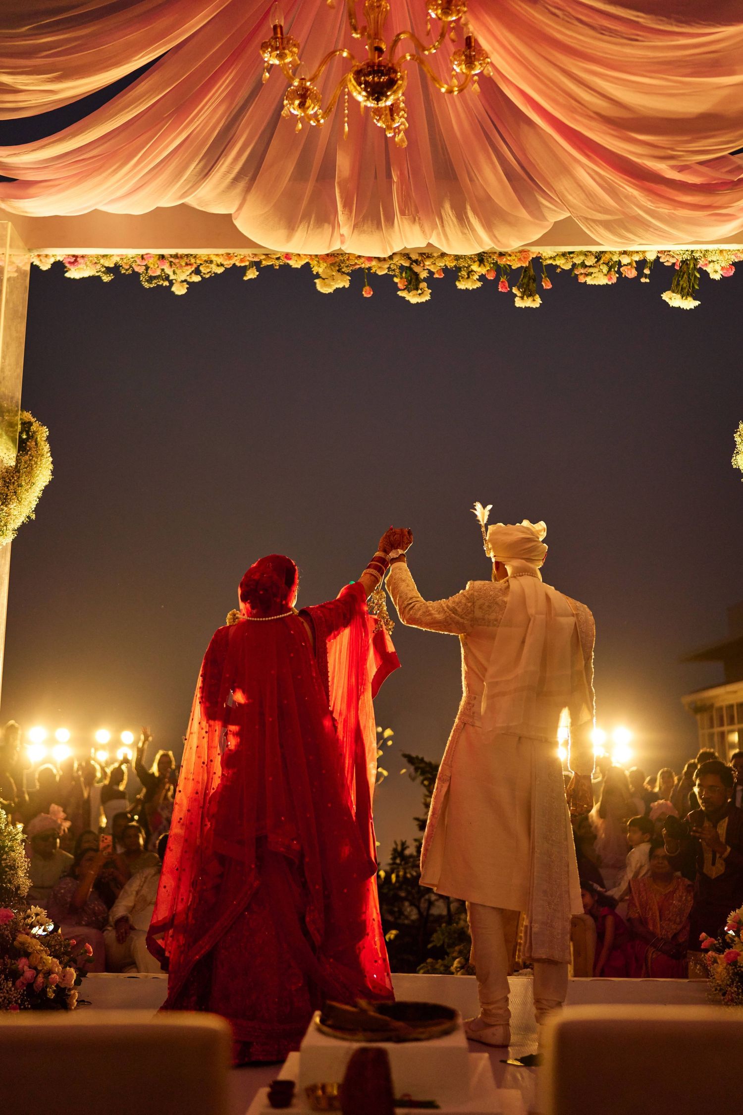 Indian bride in red lehenga and groom in white sherwani hold hands at elegant outdoor wedding ceremony at night.