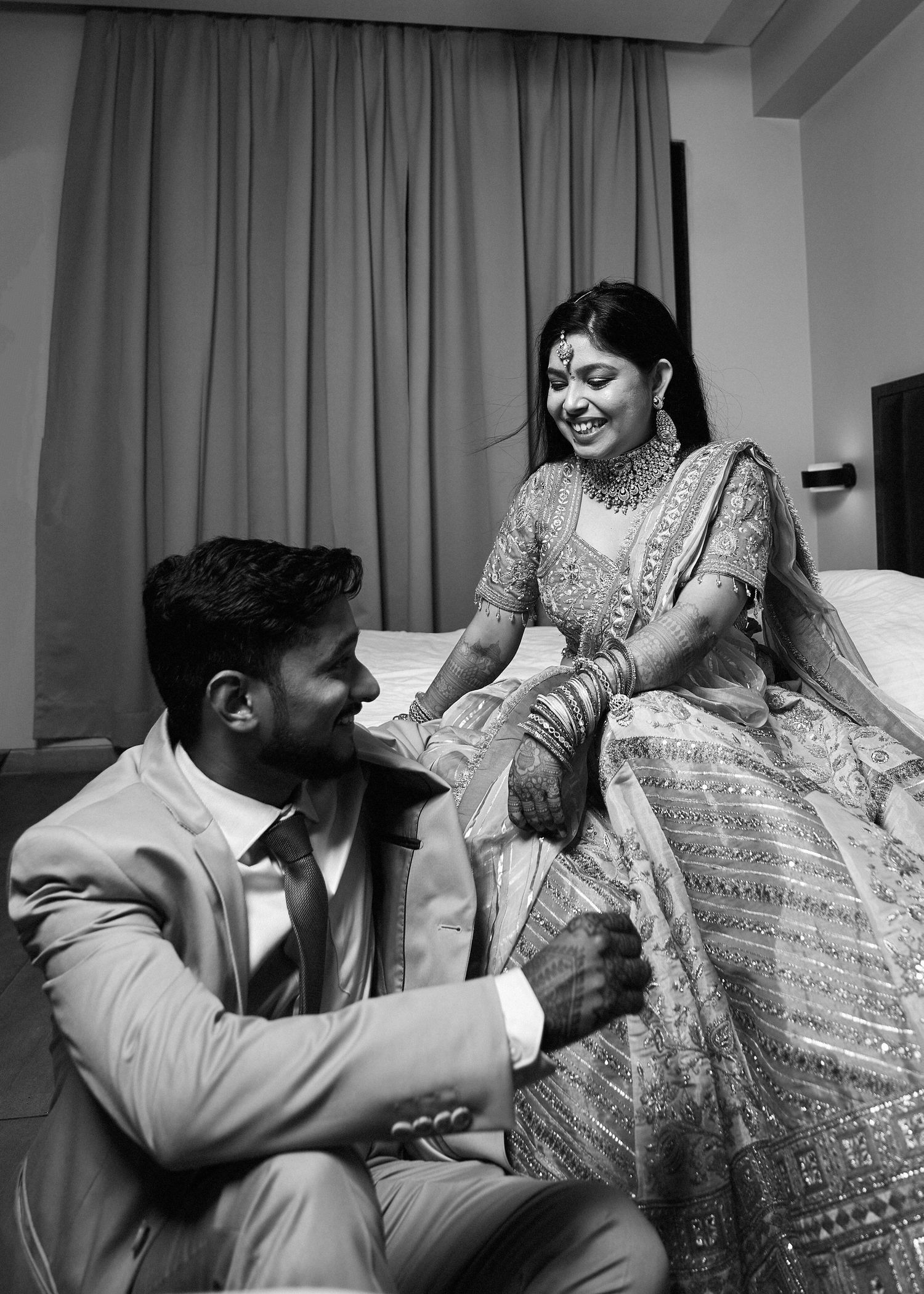 Black and white photo of an Indian bride in lehenga laughing with groom in suit during wedding celebration.