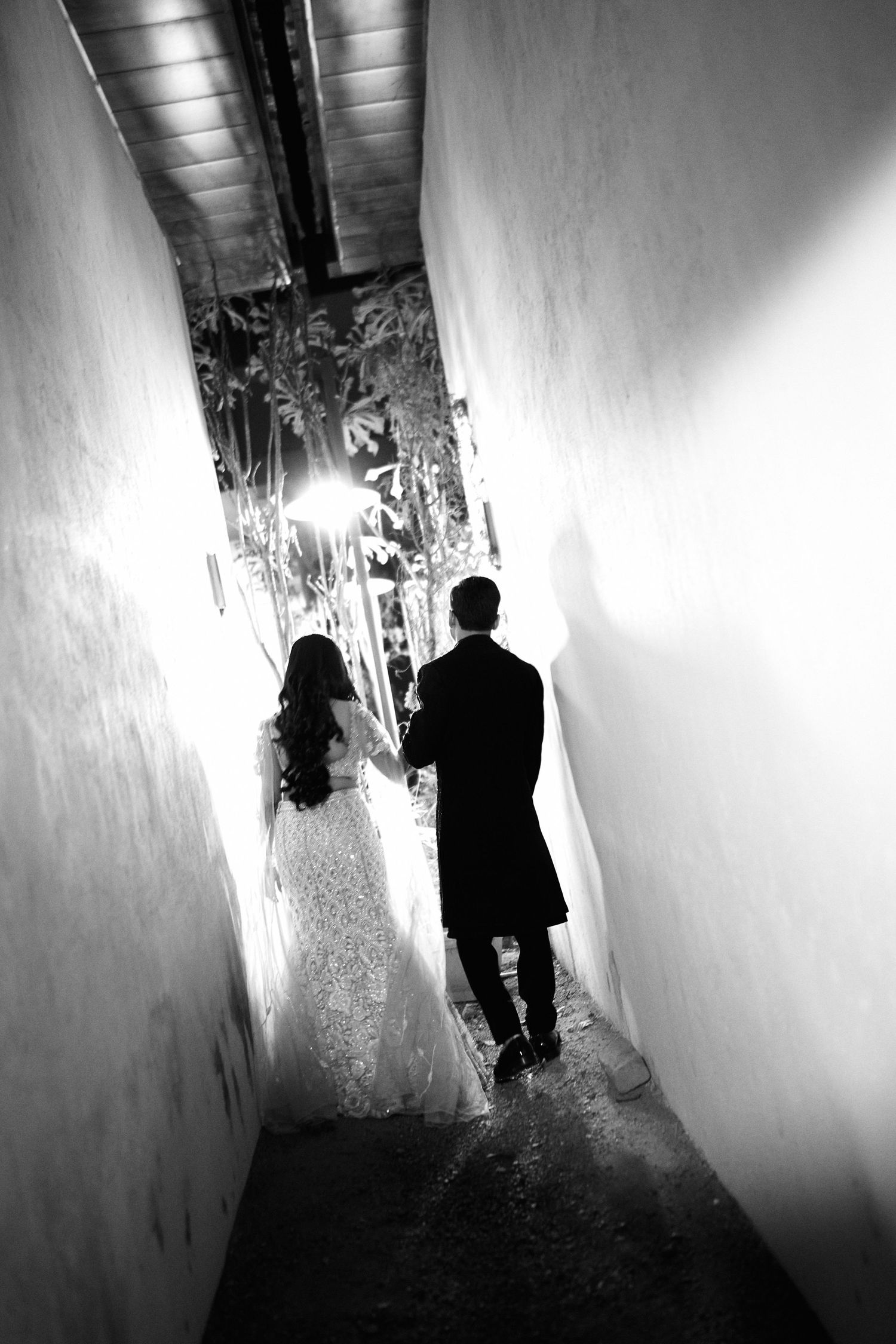 Bride and groom silhouette walking through narrow corridor toward light, black and white wedding photo.