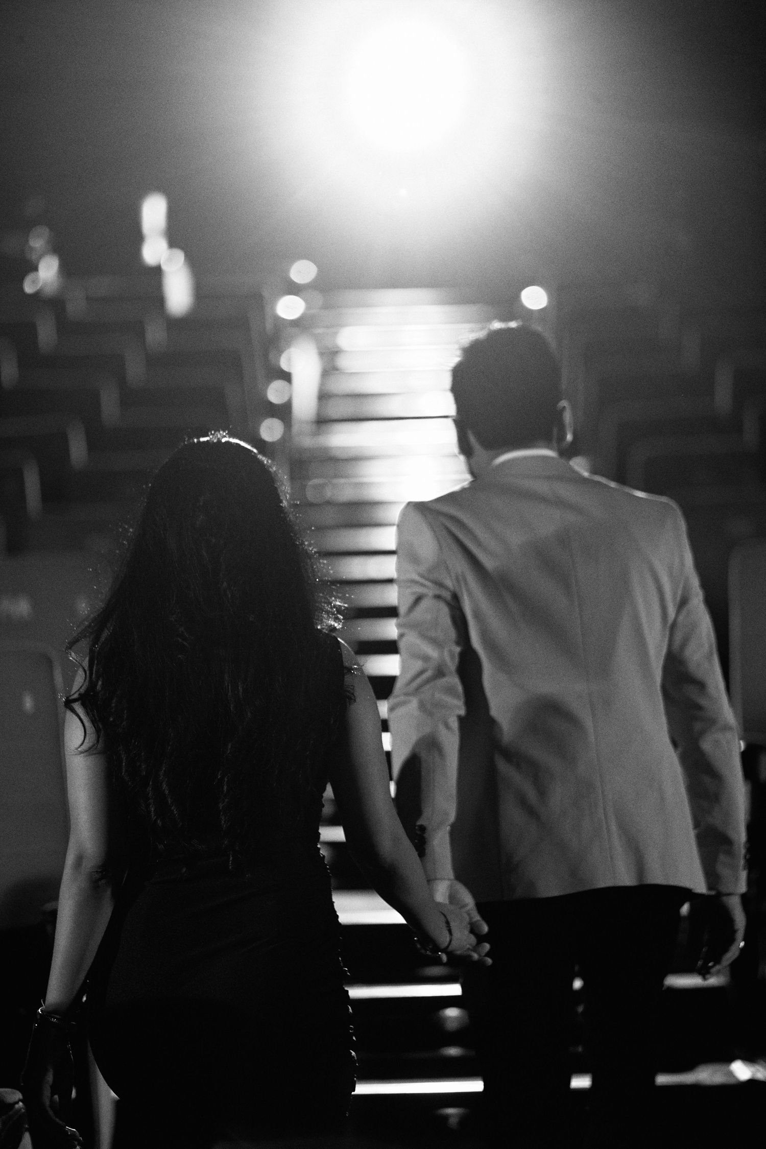 Black and white photo of a couple holding hands walking up stairs toward bright lights in a romantic setting.
