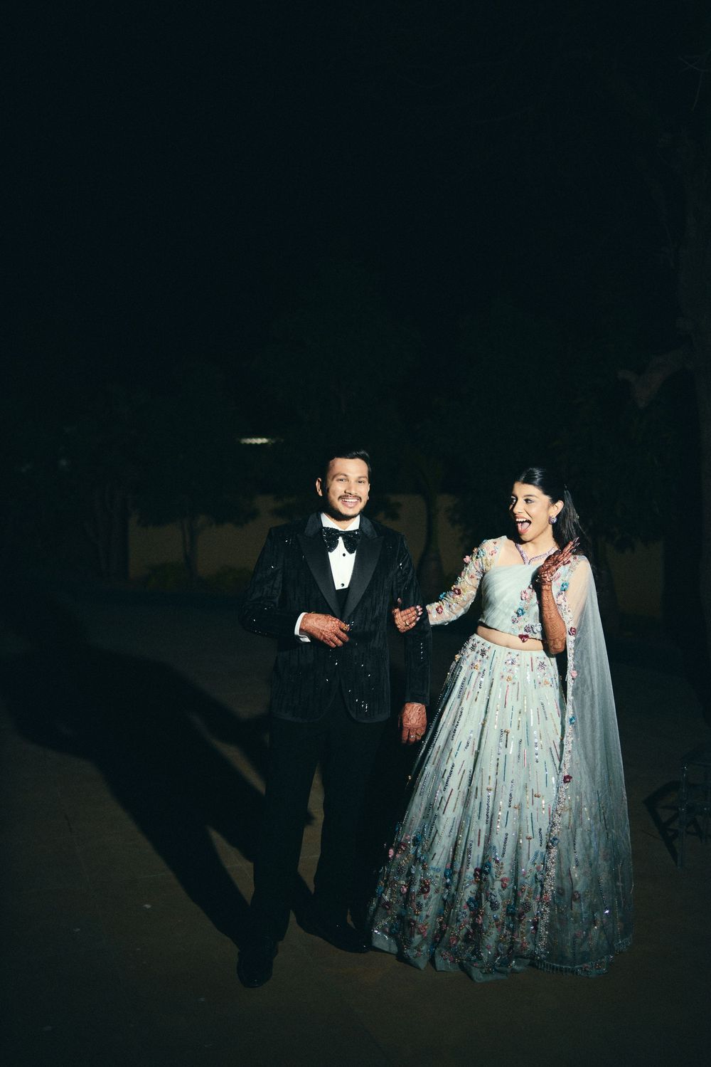 Couple in elegant wedding attire, man in black tuxedo and woman in floral lehenga, posing outdoors at night.