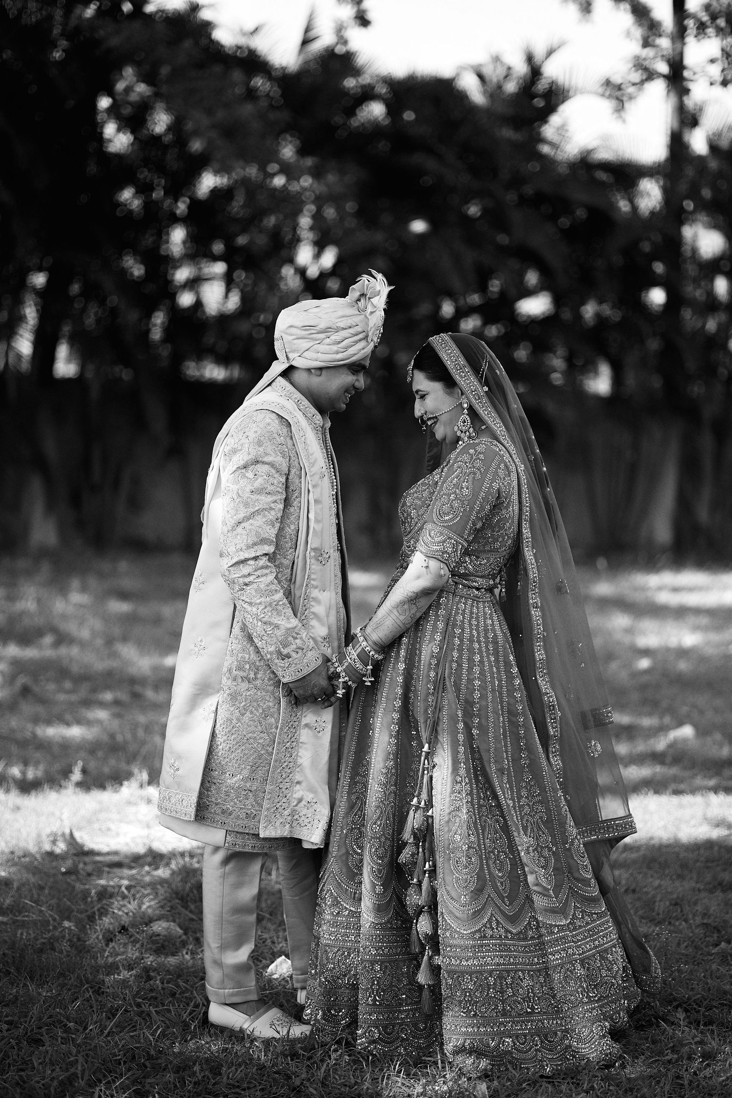Black and white photo of an Indian bride and groom in traditional attire holding hands and smiling at each other outdoors.