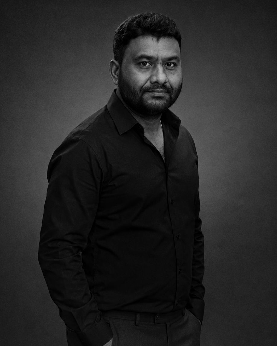 Black and white portrait of a bearded man in a black shirt posing confidently against a dark studio background.