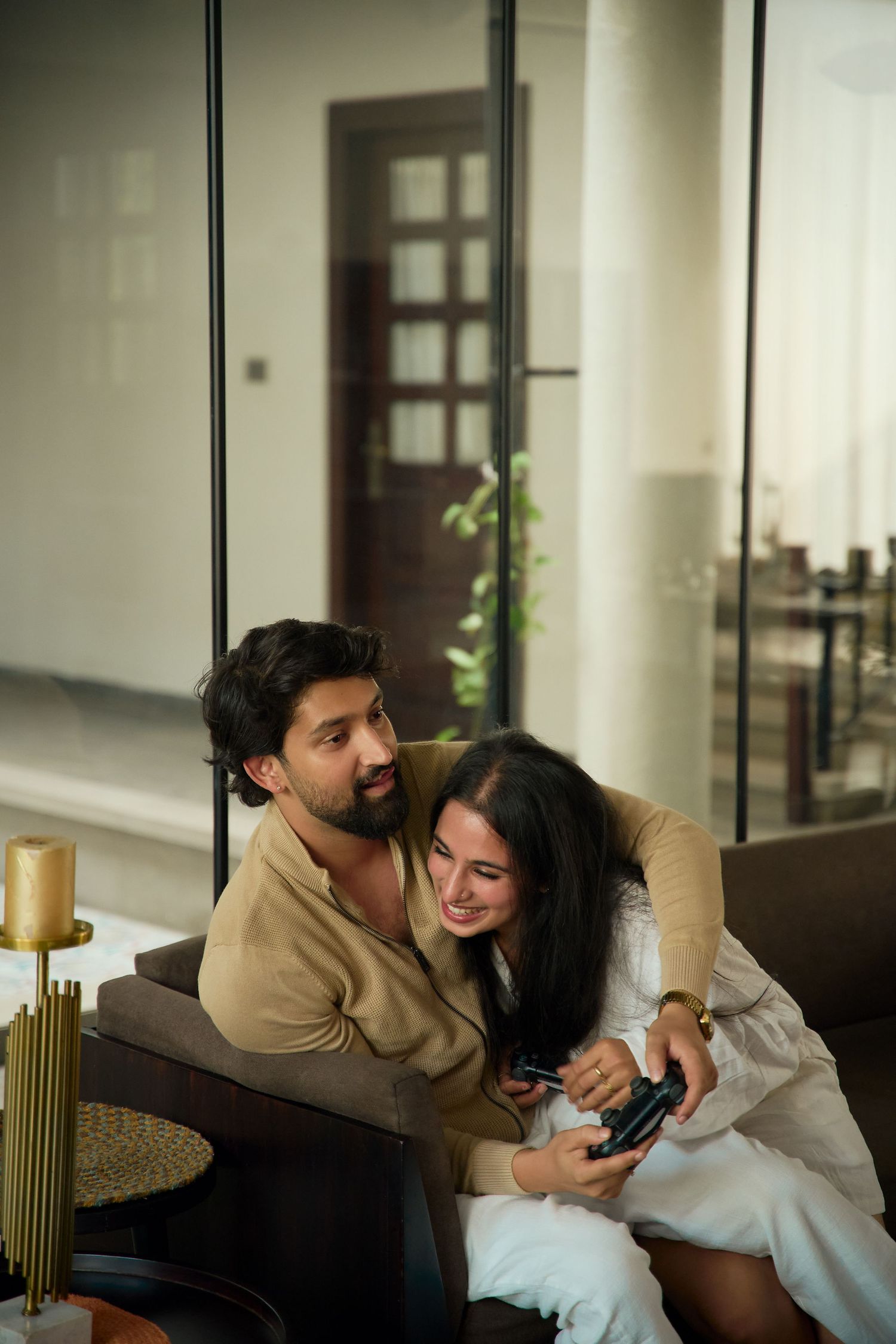 Couple sitting on sofa together, woman in white dress smiling at phone, man in tan shirt looking at her affectionately.