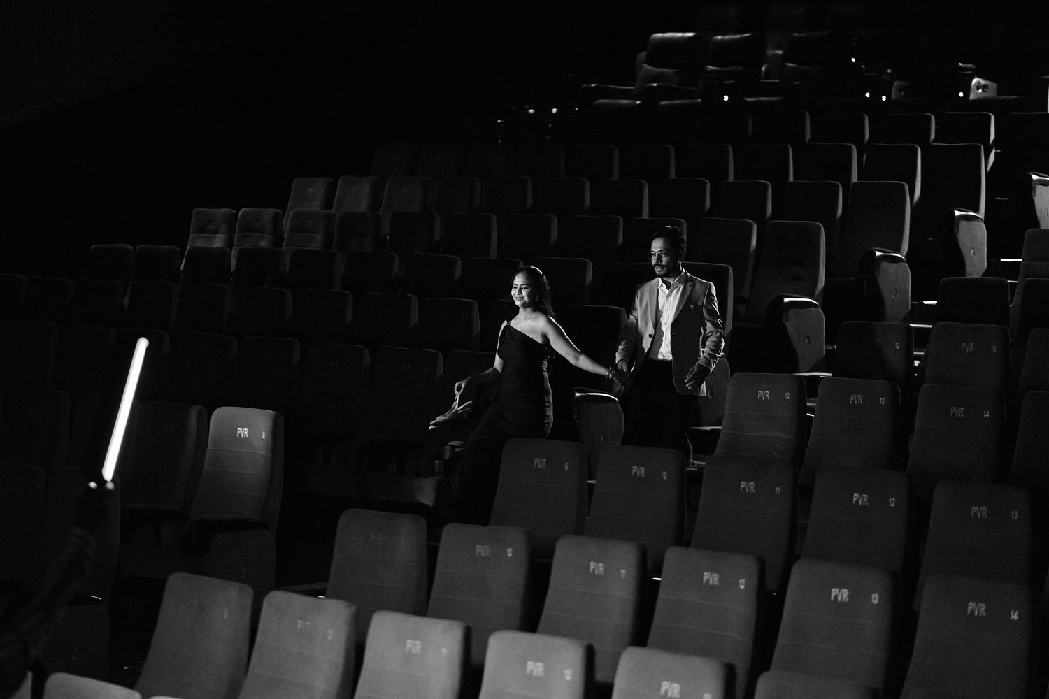 Couple holding hands walking through empty theater rows in a romantic black and white cinematic photo.