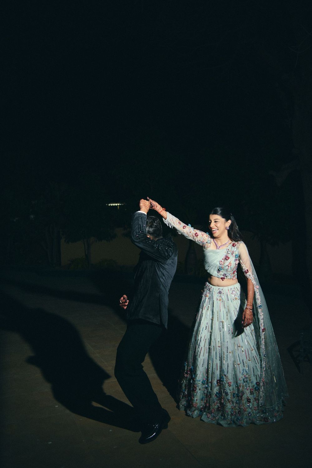 Couple dancing outdoors at night, woman in light blue floral lehenga, man in dark suit, spinning under the stars.