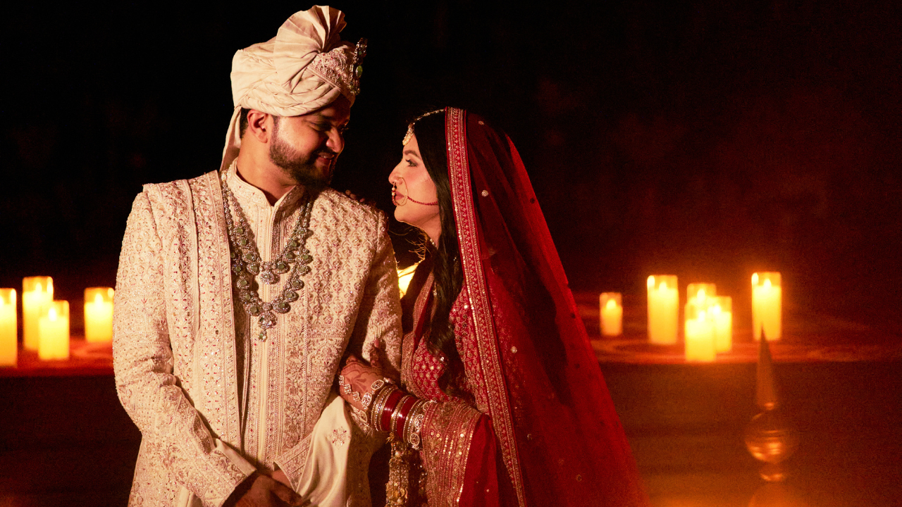 Indian bride in red lehenga and groom in cream sherwani share romantic candlelit wedding moment.