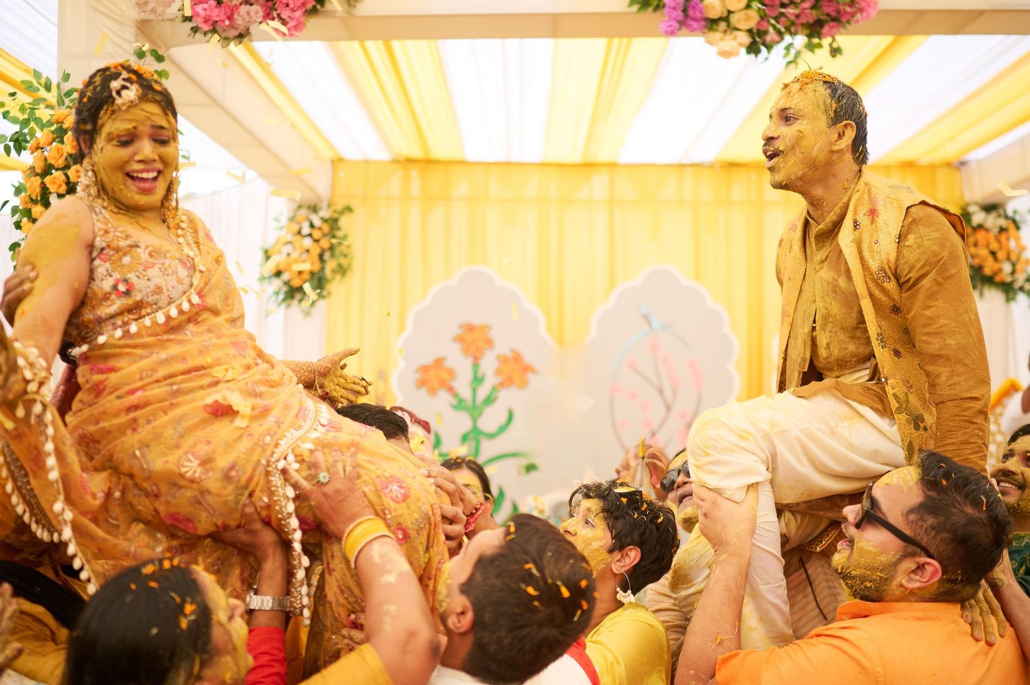 Indian couple lifted by guests during joyful Haldi wedding ceremony, dressed in traditional yellow outfits.