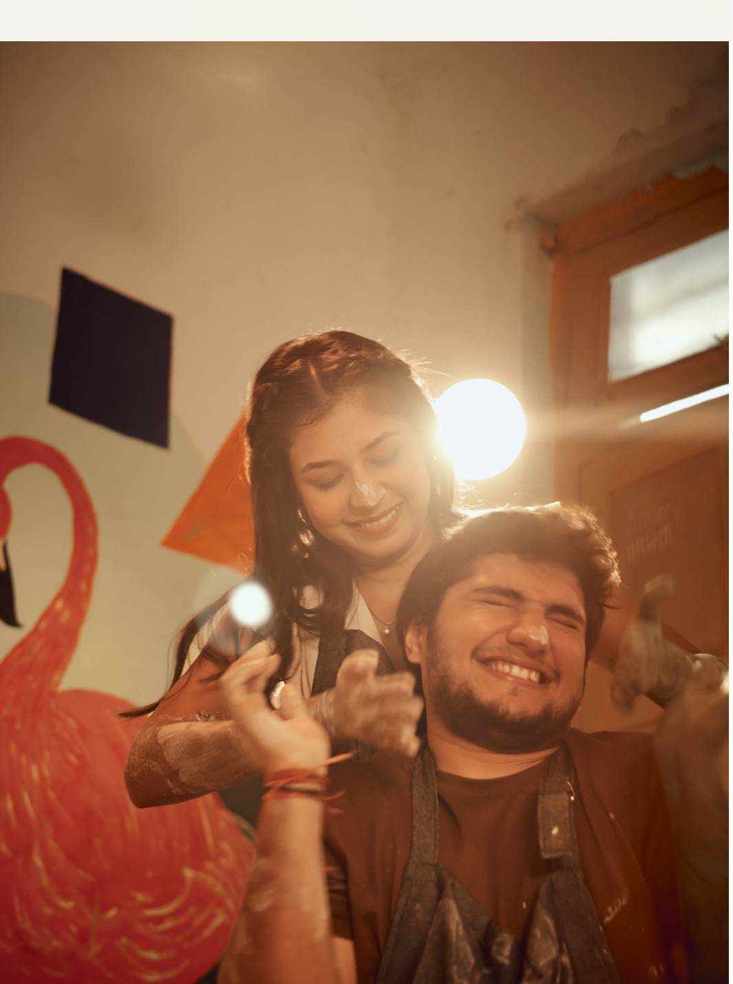 A joyful couple laughs together indoors in warm golden light, surrounded by colorful wall decorations.