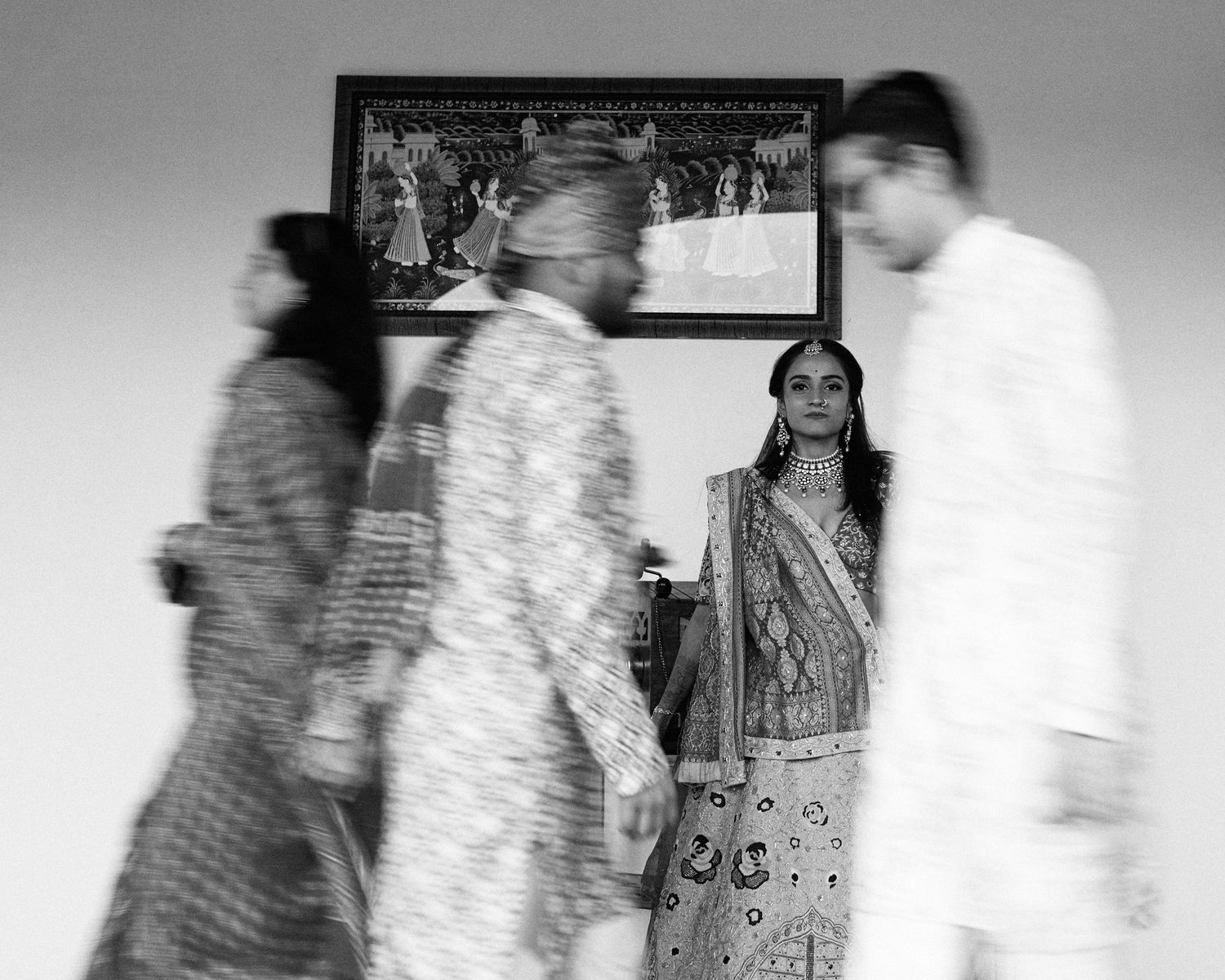 Black and white photo of people in traditional Indian attire, with a woman in embroidered lehenga standing still amid motion.
