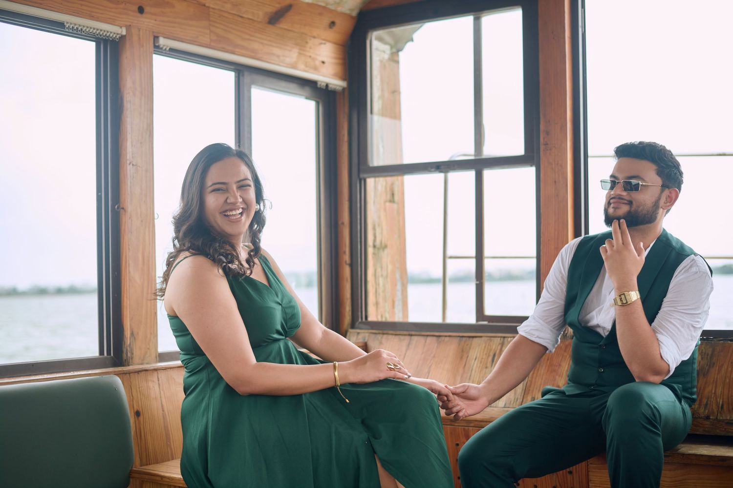Couple in green outfits sitting together in a wooden lakeside cabin, smiling and holding hands.