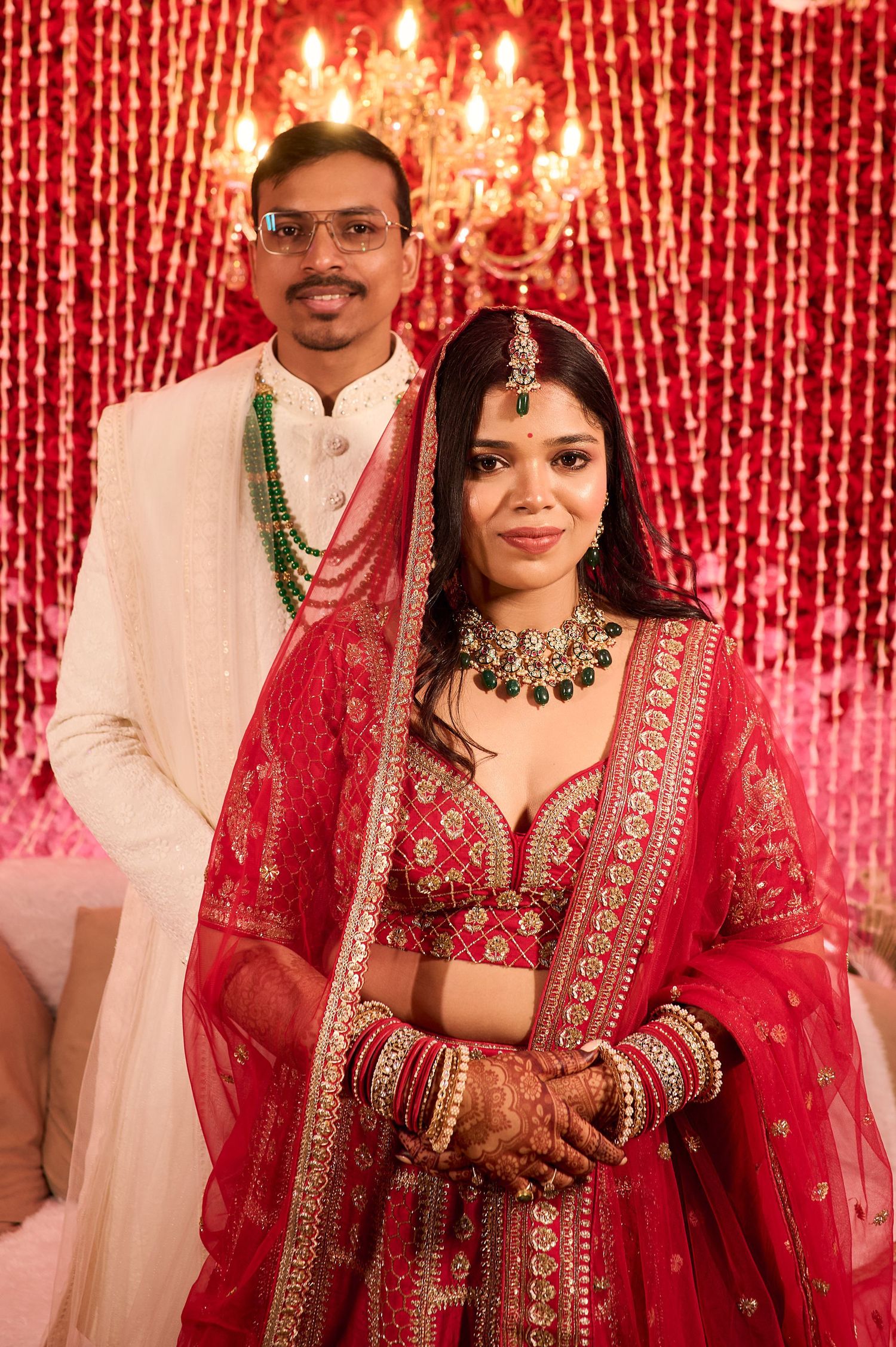 Indian bride in red lehenga with green jewelry stands with groom in white sherwani at traditional wedding ceremony.