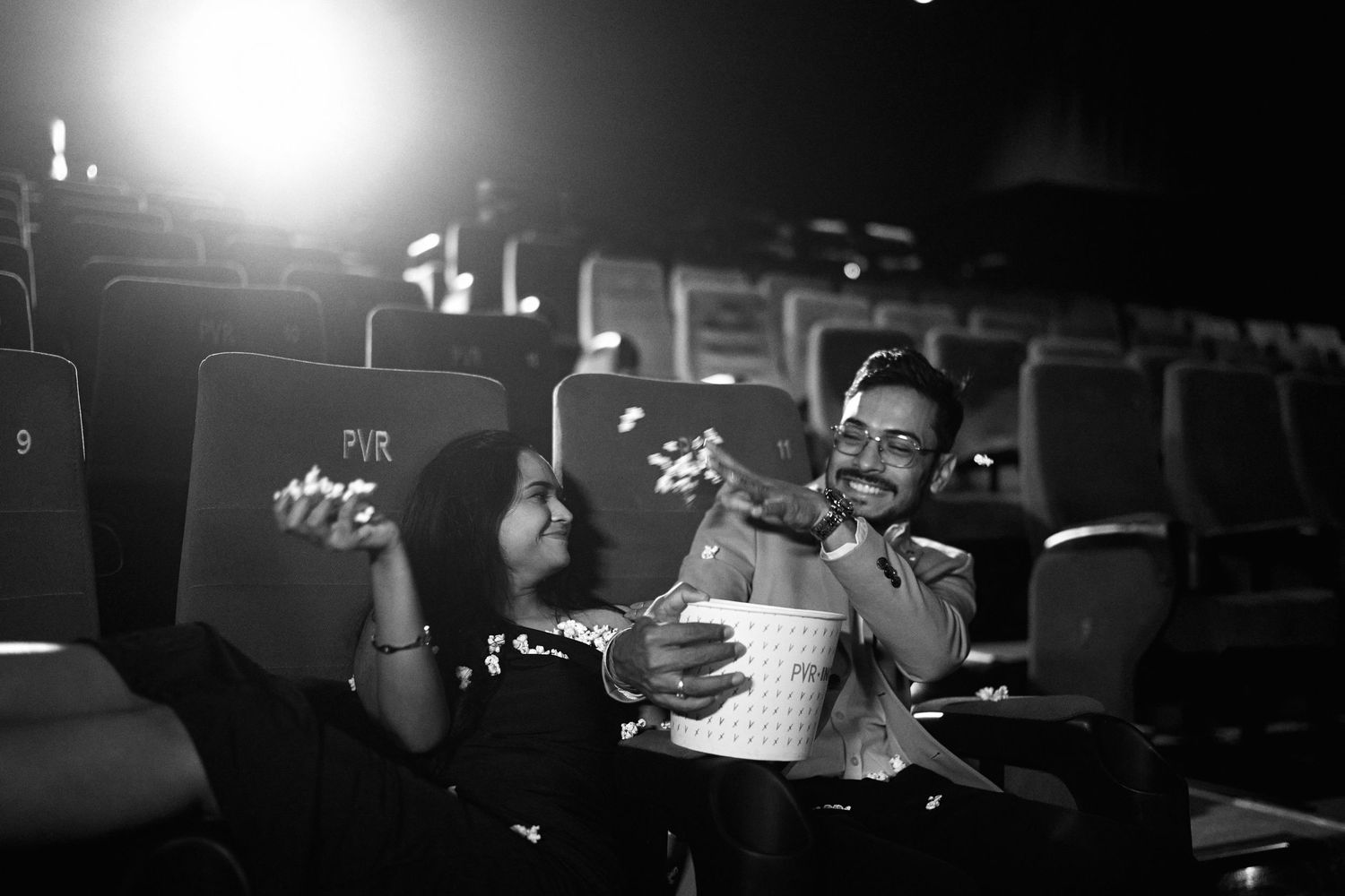 Black and white photo of a couple sharing popcorn and laughing together in a movie theater.