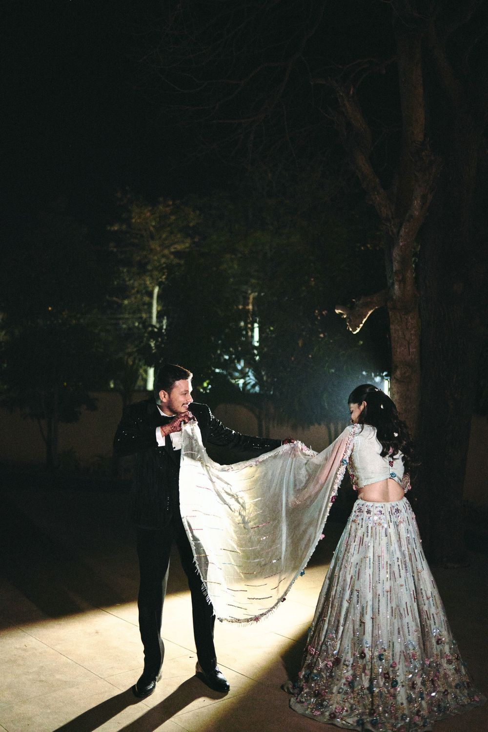 Couple sharing a romantic dance outdoors at night, bride in floral lehenga, groom in black tuxedo, veil flowing gracefully.
