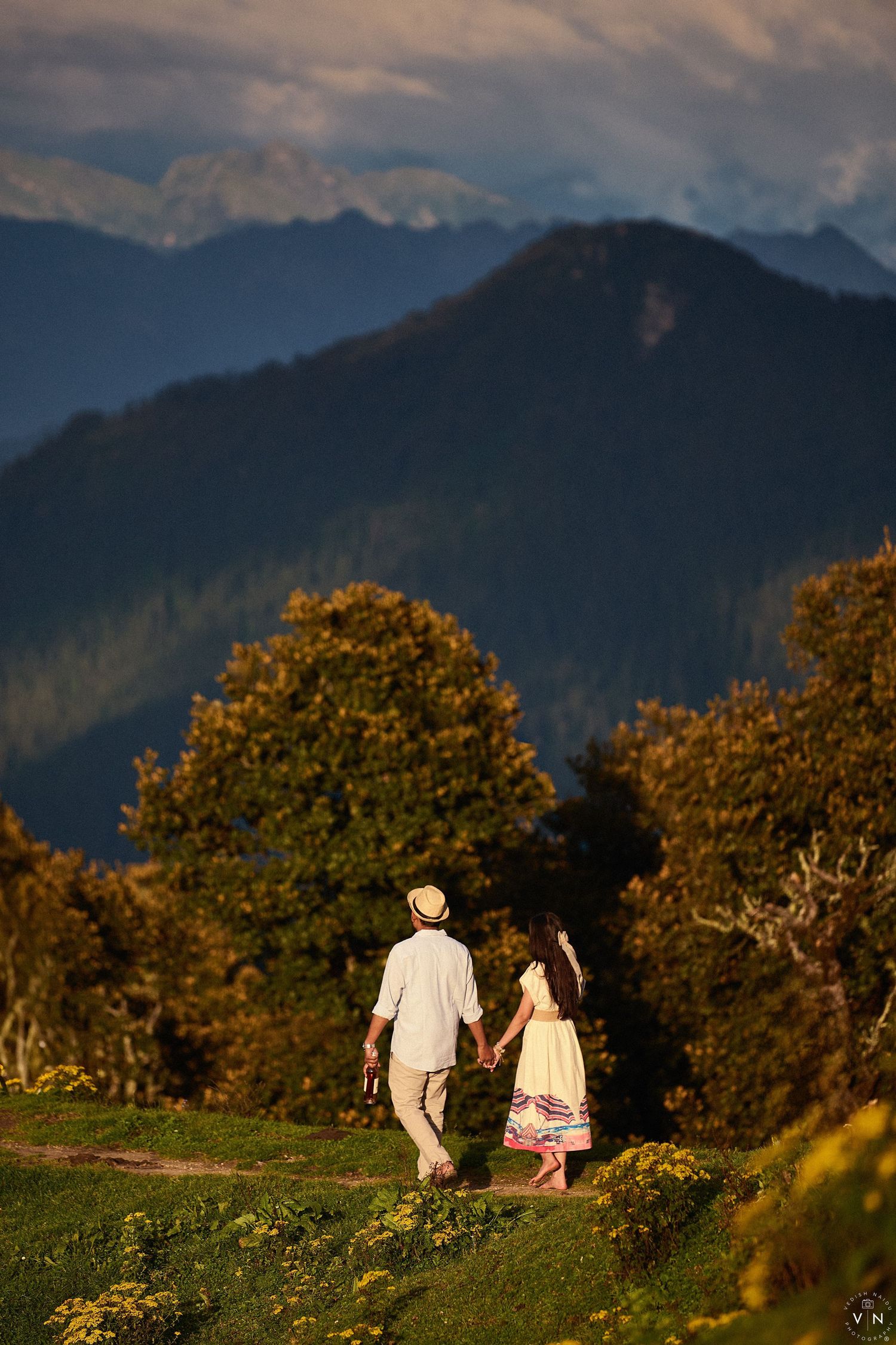 Couple holding hands walking through green meadow with mountain landscape at golden hour.
