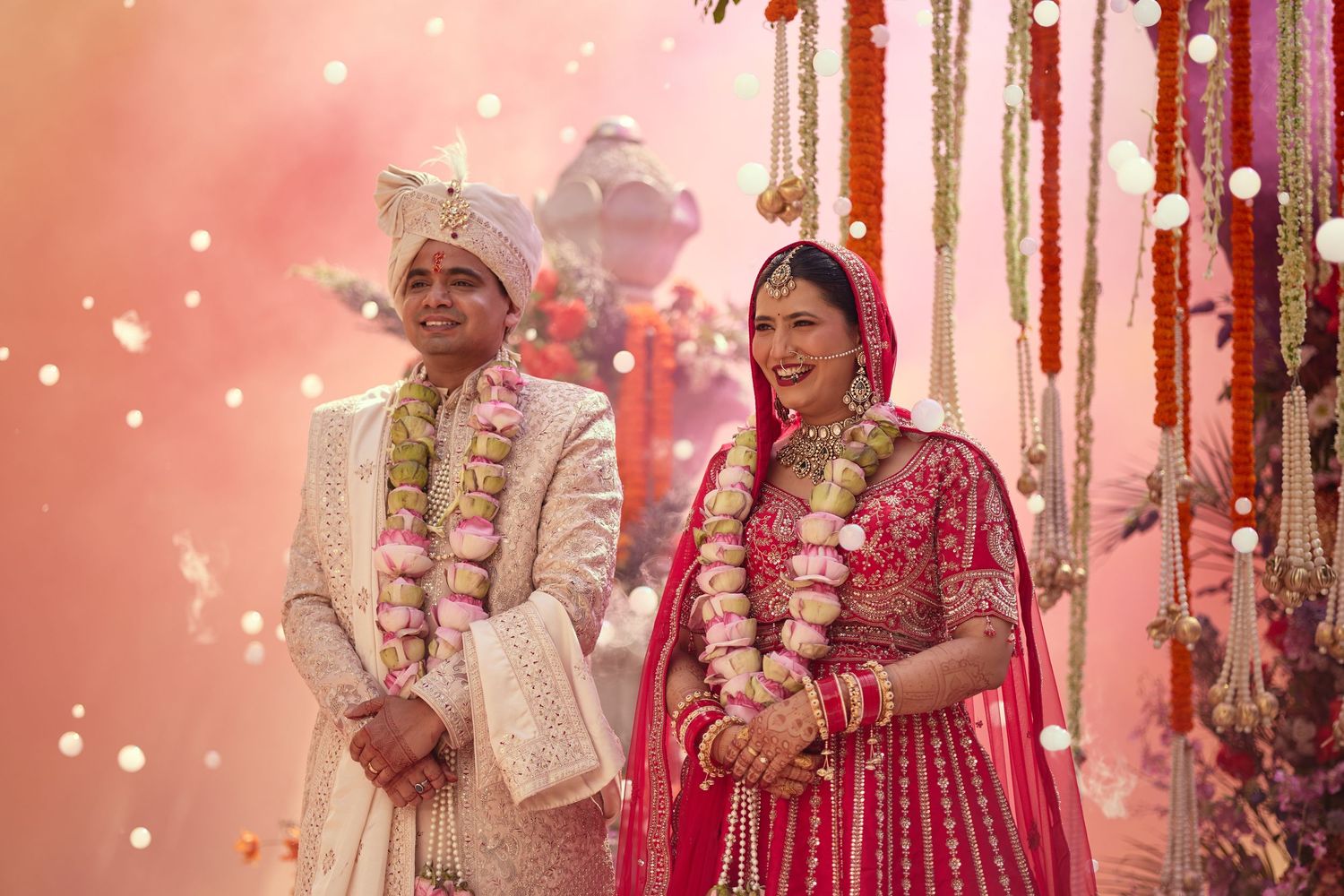Joyful Indian bride in pink lehenga and groom in cream sherwani celebrate wedding with floral garlands.