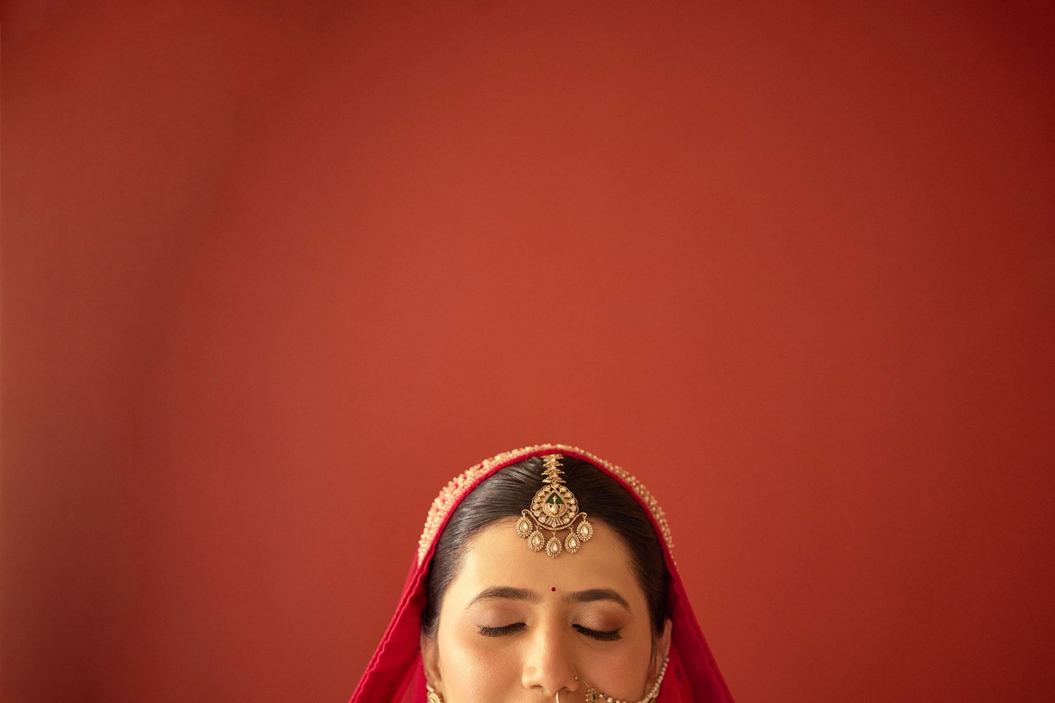 Indian bride wearing red veil and gold maang tikka with eyes closed against red wall.