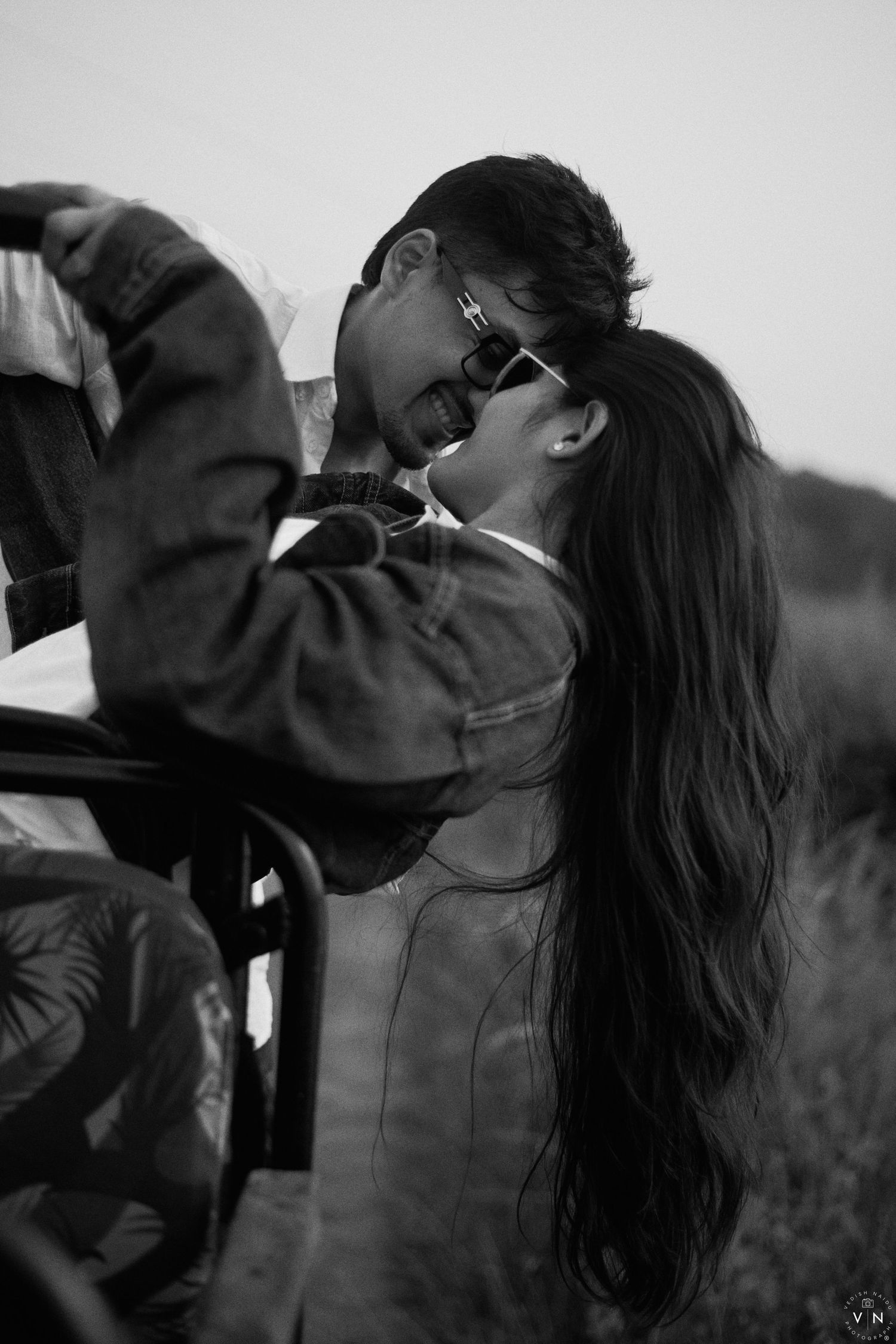 Black and white photo of a romantic couple sharing an intimate moment, woman with long hair and sunglasses leaning back.