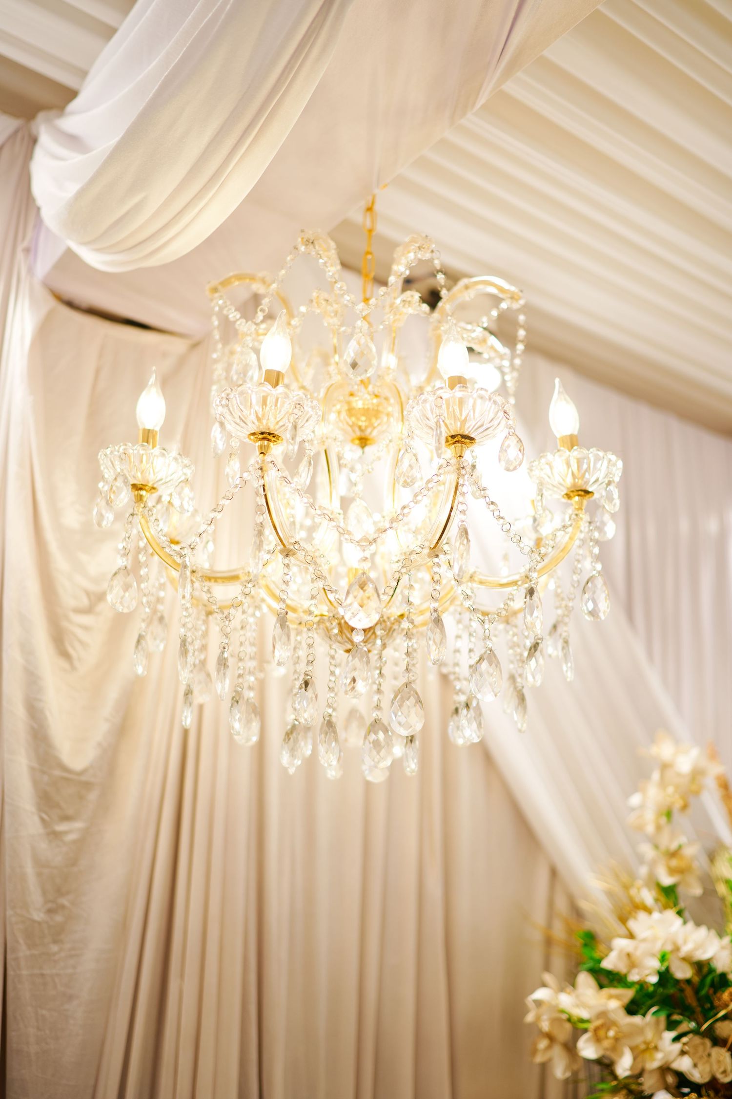 Elegant crystal chandelier with gold accents hanging from draped white fabric ceiling at a wedding venue.