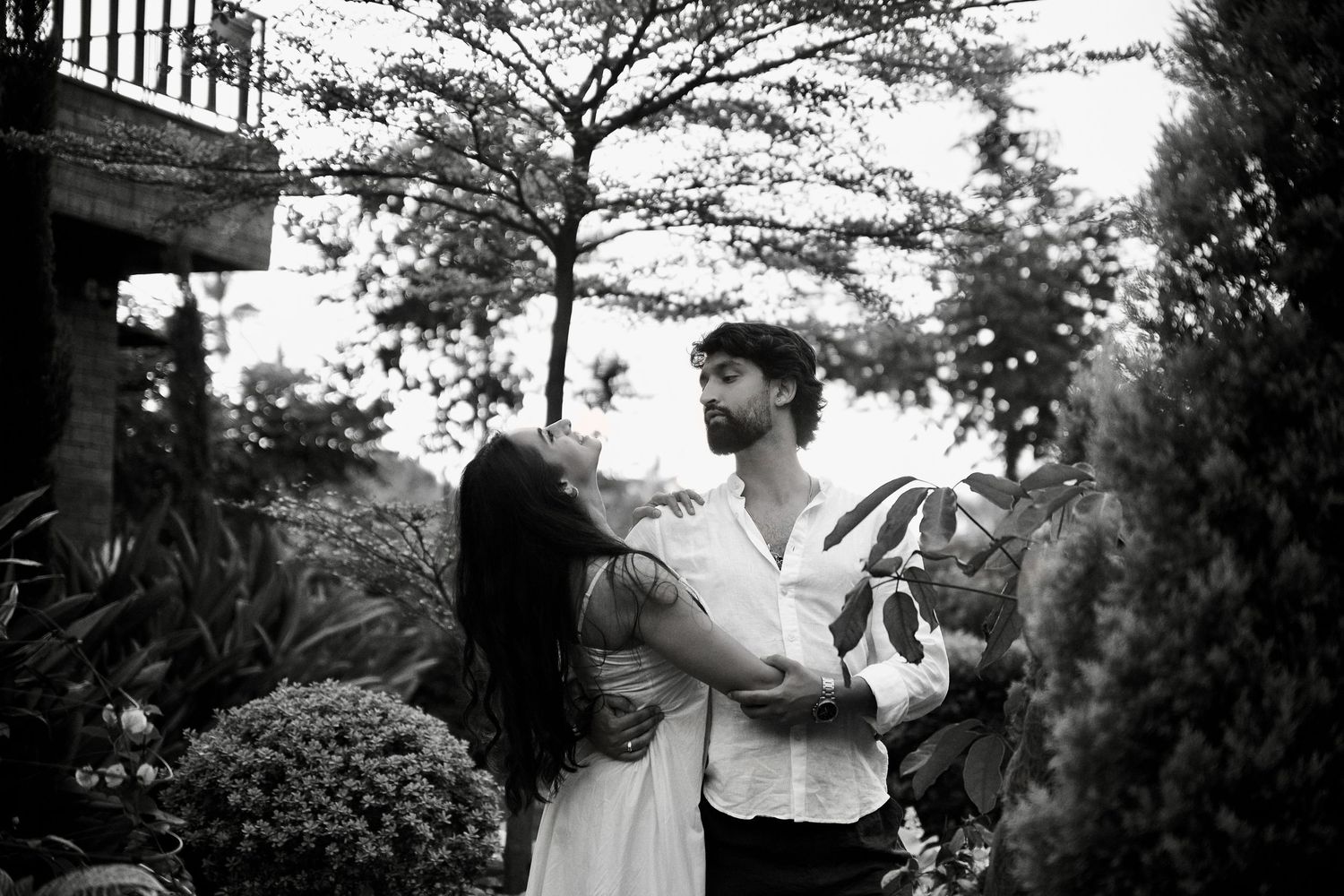 Black and white photo of a couple embracing in a lush garden, man holding a small dog while woman touches his face.