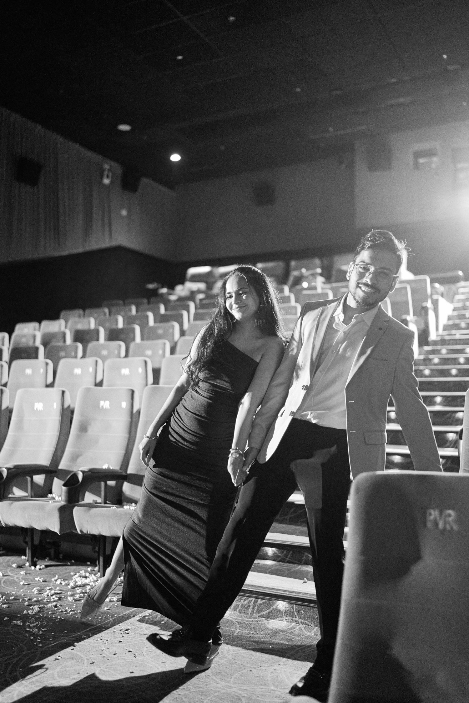 Elegant couple in formal attire posing in empty PVR cinema hall, black and white photography.