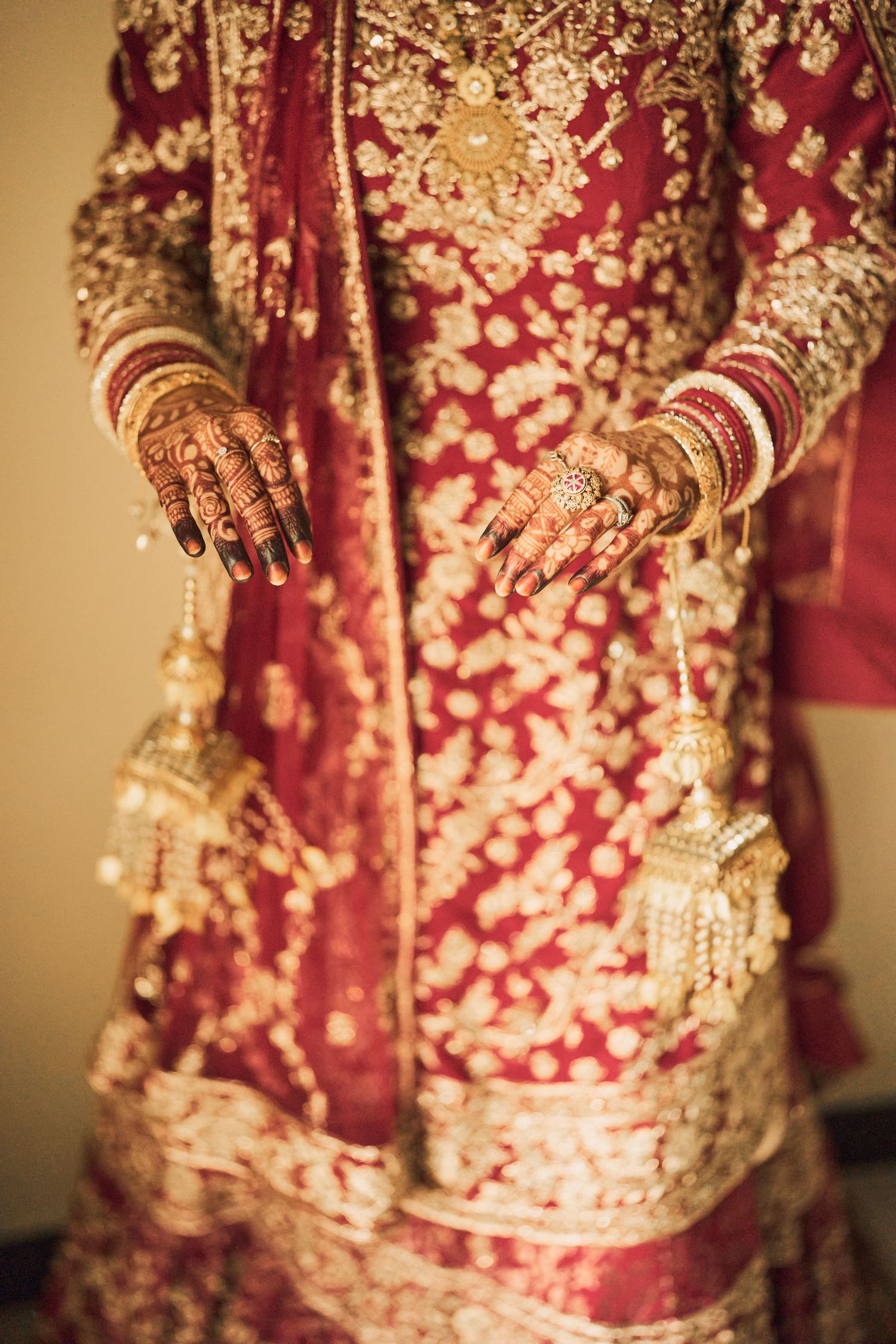 Indian bride in red and gold lehenga with henna hands and gold bangles at wedding ceremony.